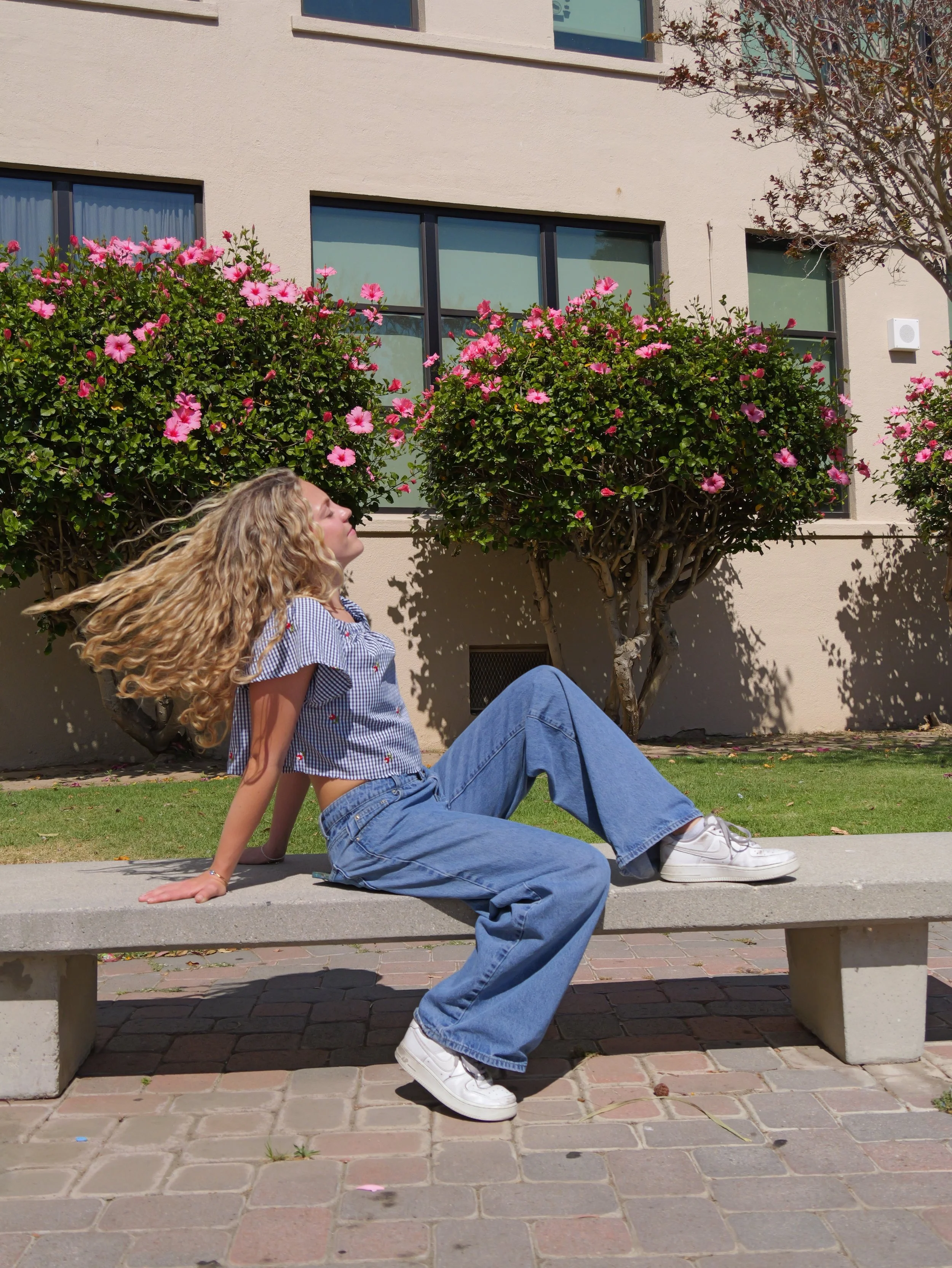 A young woman with curly blonde hair sits on a park bench, leaning back with her eyes closed, enjoying the sunshine. She wears a blue and white checkered shirt, blue jeans, and white sneakers. Behind her is a beige building with large windows and pin