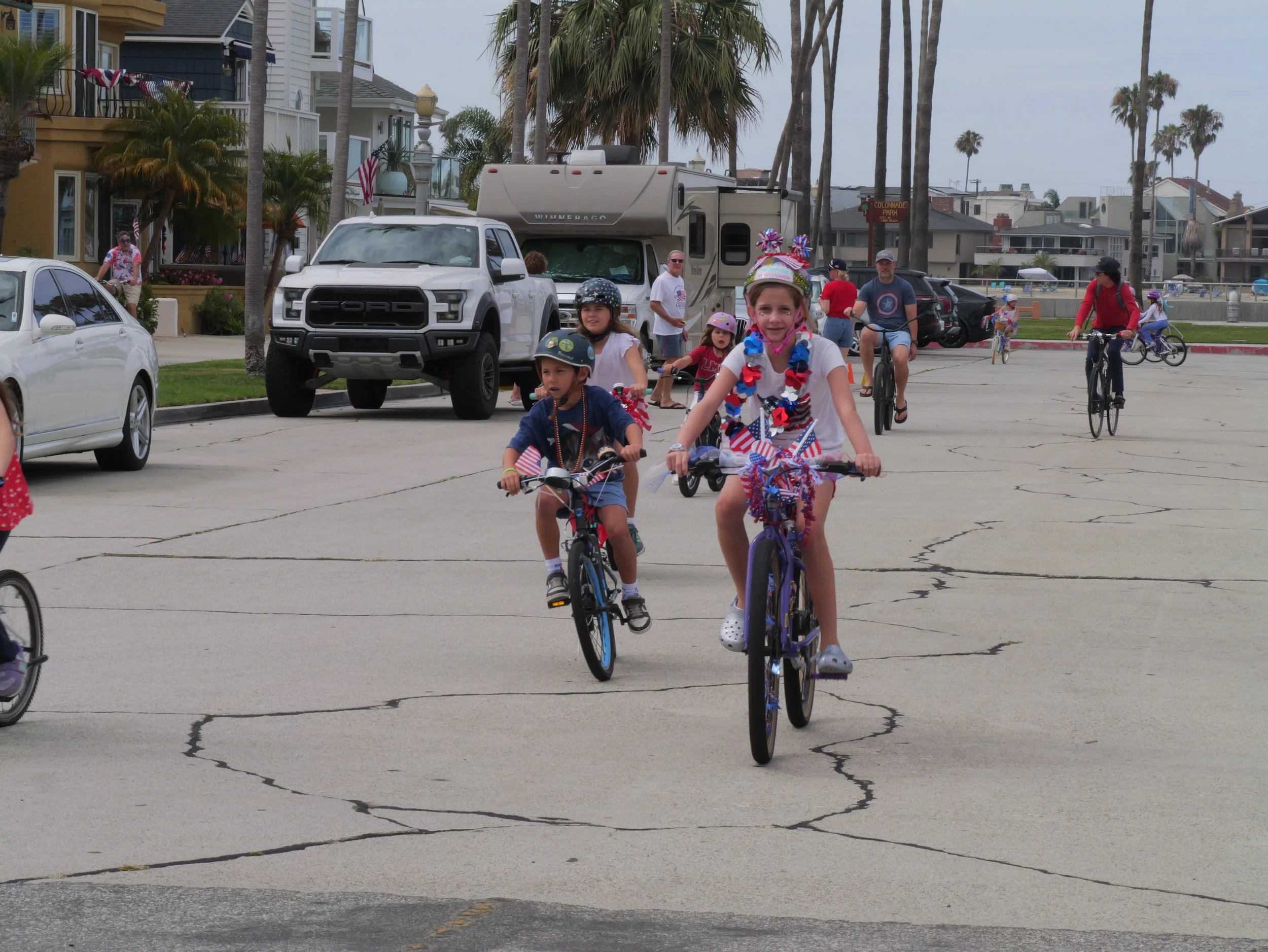 Children and adults riding bikes in a parade with festive decorations in a residential area with palm trees and houses.