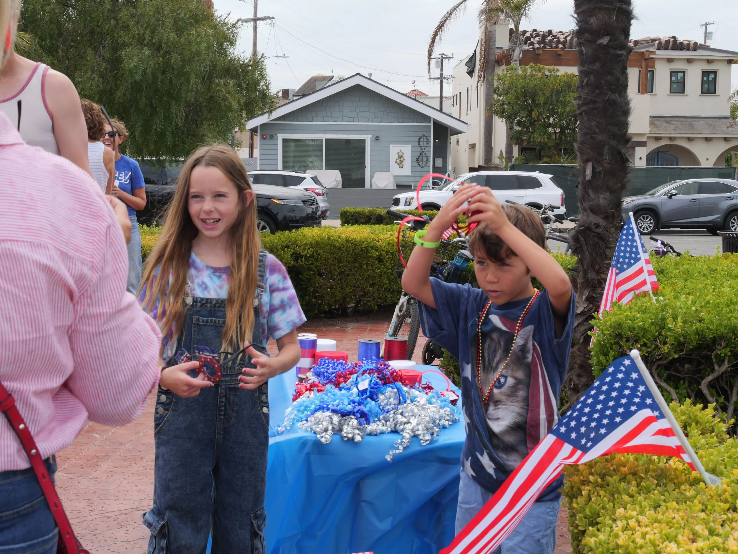 Children gathered around a table with red, white, and blue patriotic decorations, celebrating outdoors near houses and parked cars, with American flags visible.