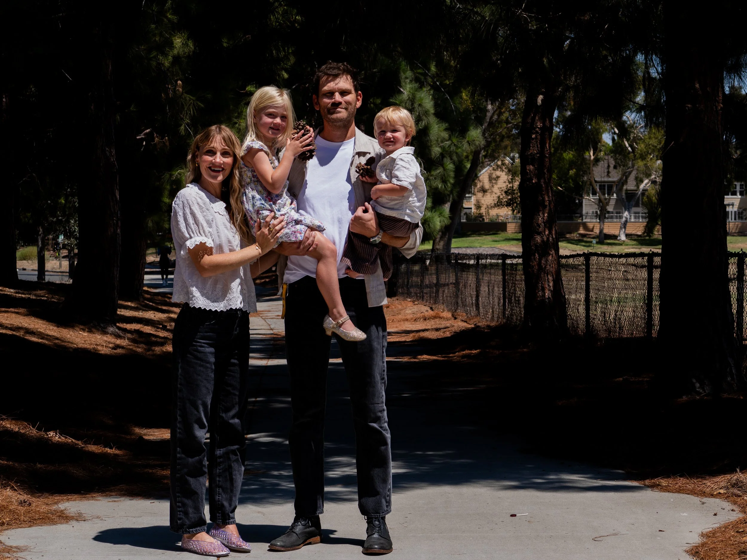 A family of five standing outdoors on a sunny day with trees and residential buildings in the background. The adults are smiling, one child is sitting on the adult's arm, and others are standing close, holding pinecones.