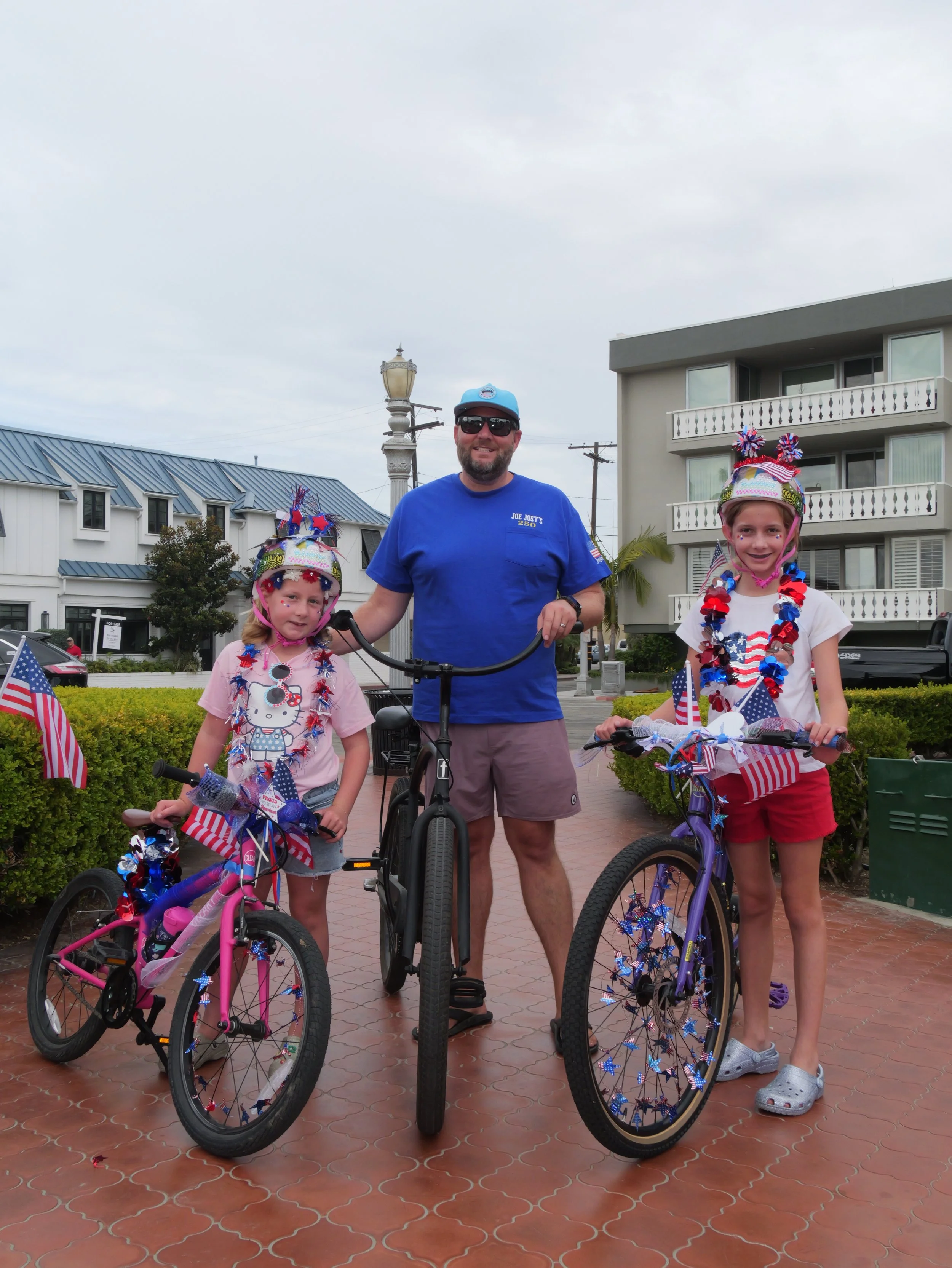 A man and two young girls with bicycles decorated with patriotic American flags and star-shaped decorations, standing outdoors on a brick-paved area, celebrating a patriotic holiday.