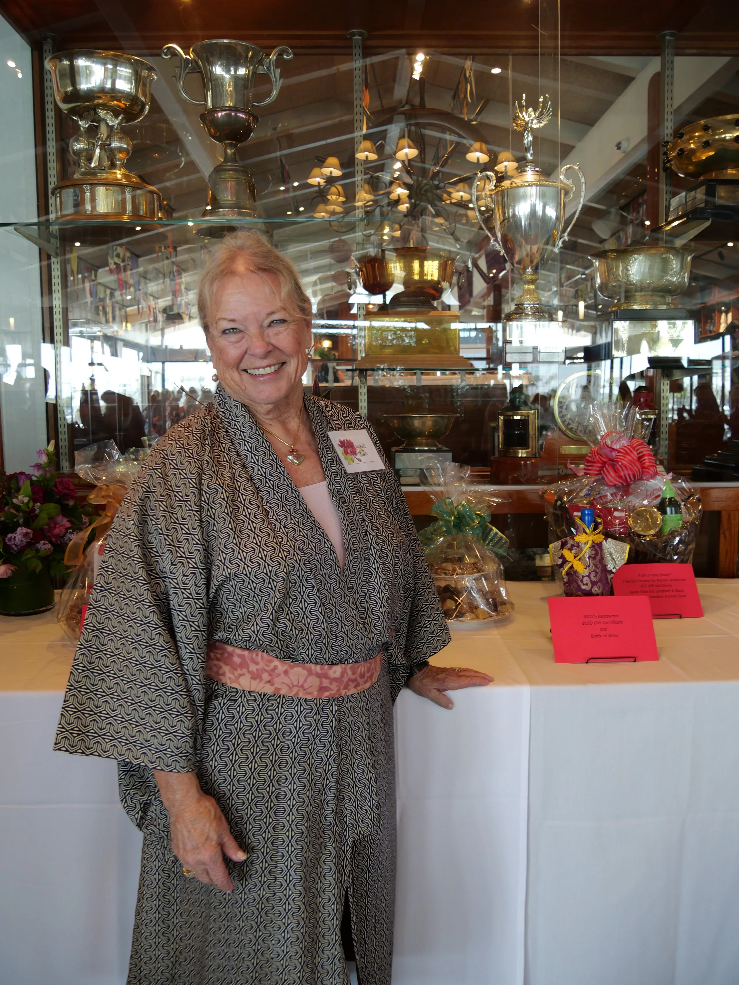 A smiling elderly woman in a patterned dress stands in front of a display case with trophies and gift baskets, in an indoor setting with warm lighting and a chandelier above.