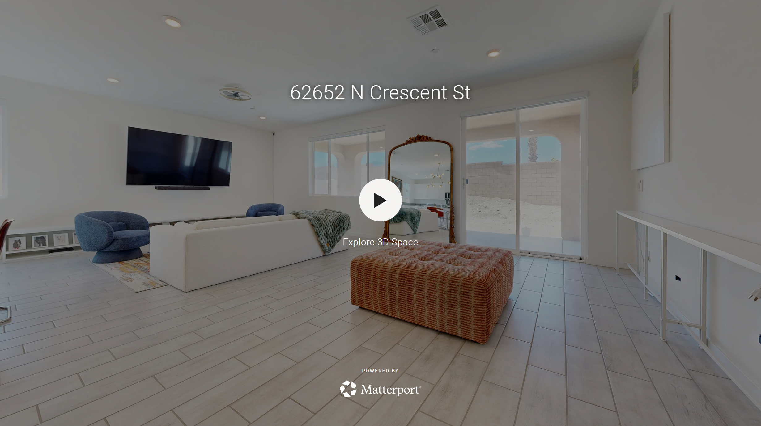 Living room with white walls, light-colored floor tiles, a large flat-screen TV on the wall, a white sofa, a blue armchair, a patterned ottoman, and large sliding glass doors leading outside.