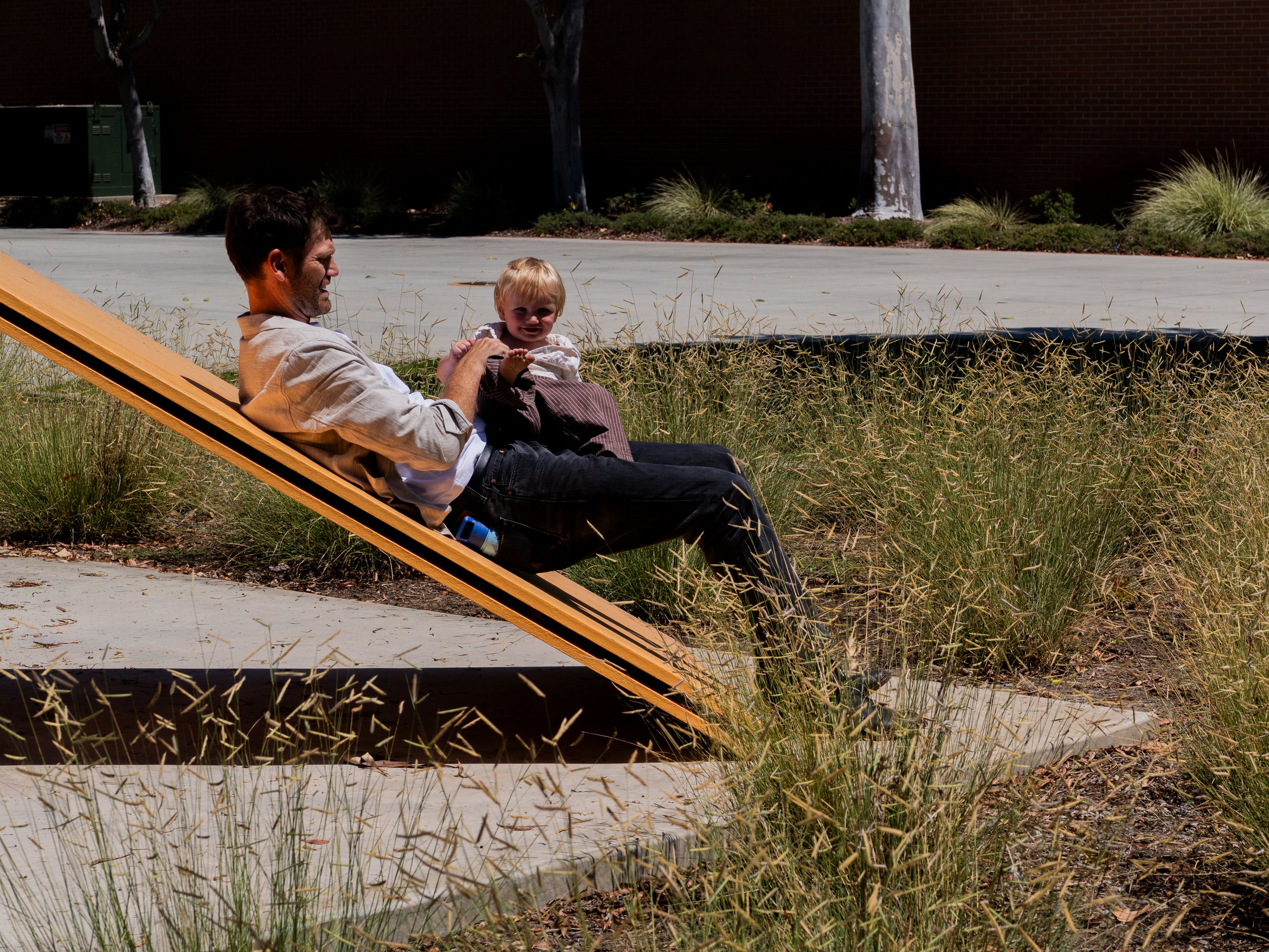 A man and a young girl sitting on a wooden deck chair outdoors. They are smiling and enjoying each other's company, with grass and trees in the background.
