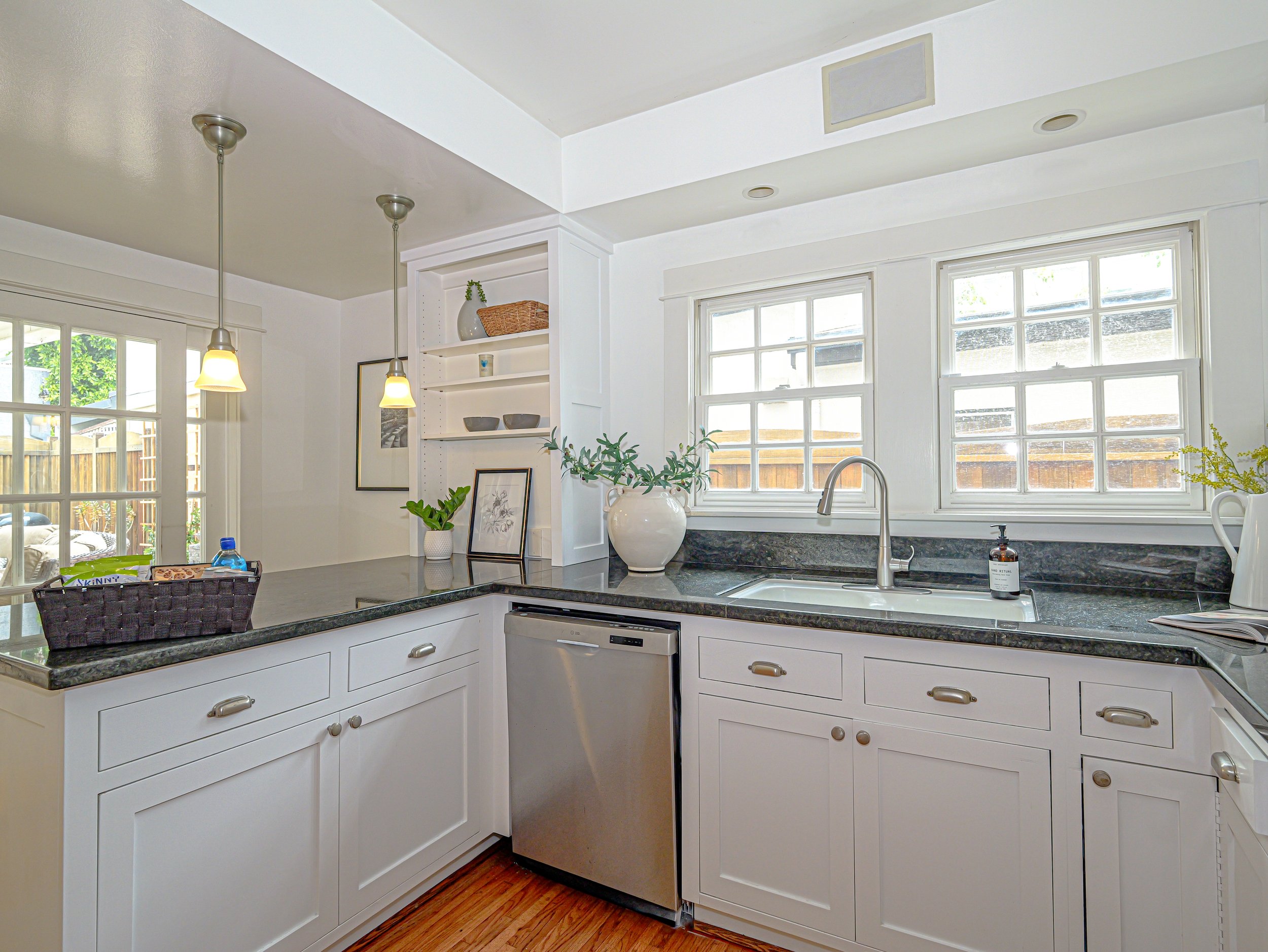 A kitchen with white cabinets, a black granite countertop, multiple windows, and decorative items including framed pictures and potted plants.