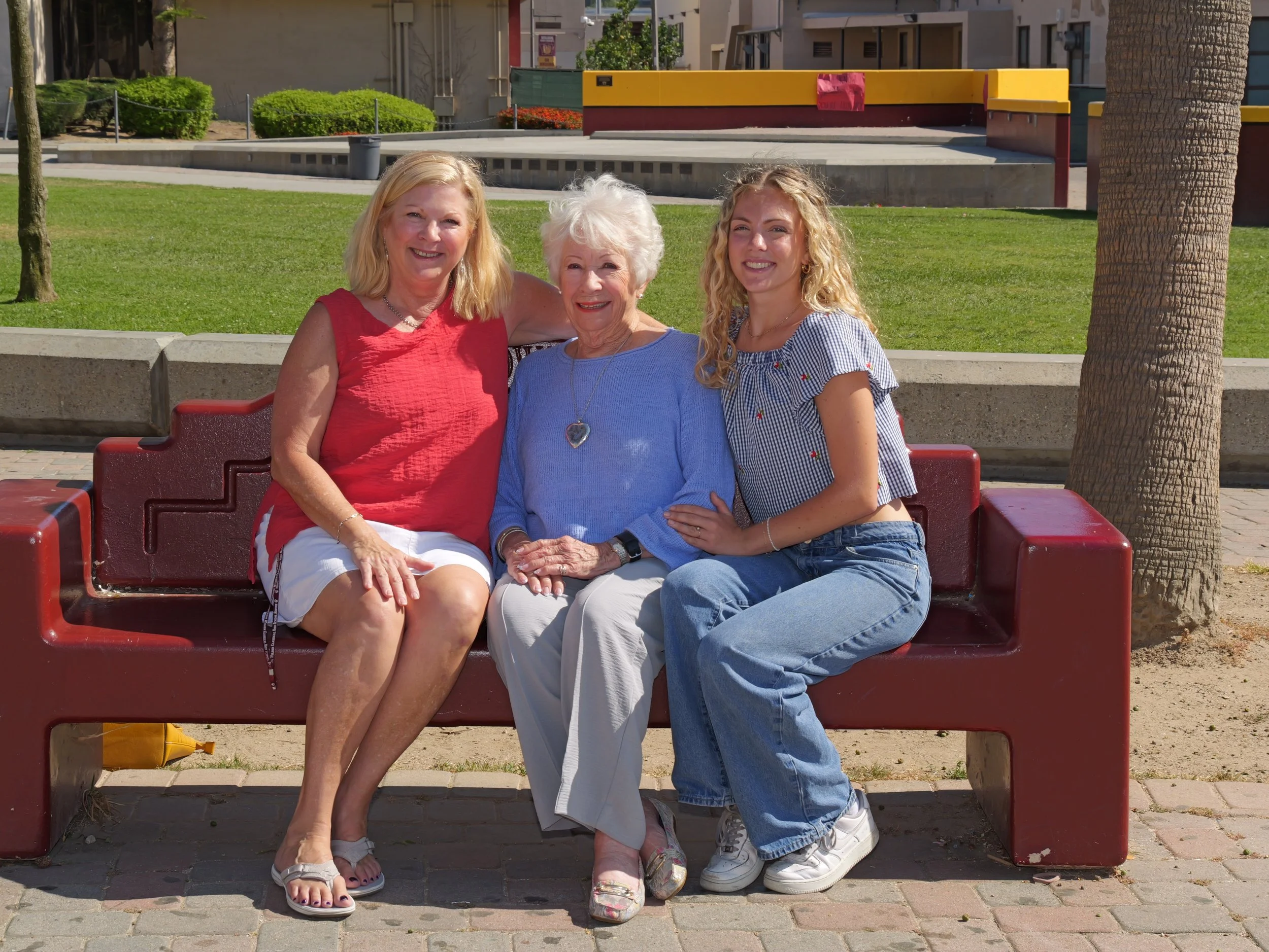 Three women sitting on a red park bench outdoors on a sunny day, smiling at the camera. The woman on the left has blonde hair and is wearing a red sleeveless top and white shorts. The woman in the middle has short white hair and is dressed in a blue 