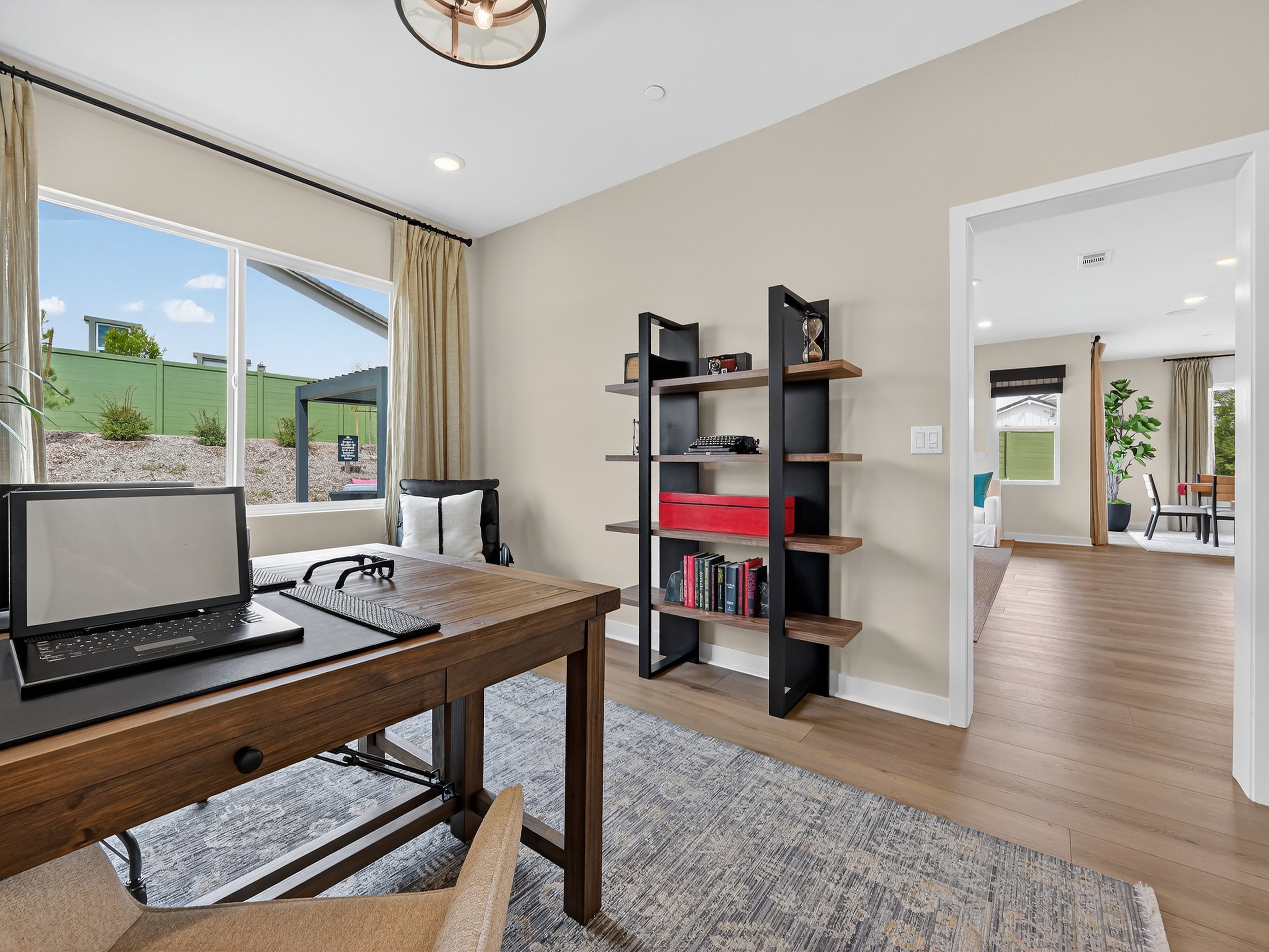 Interior of a modern living room with large window, beige walls, kitchen at the back, bookshelf, and wooden floor.