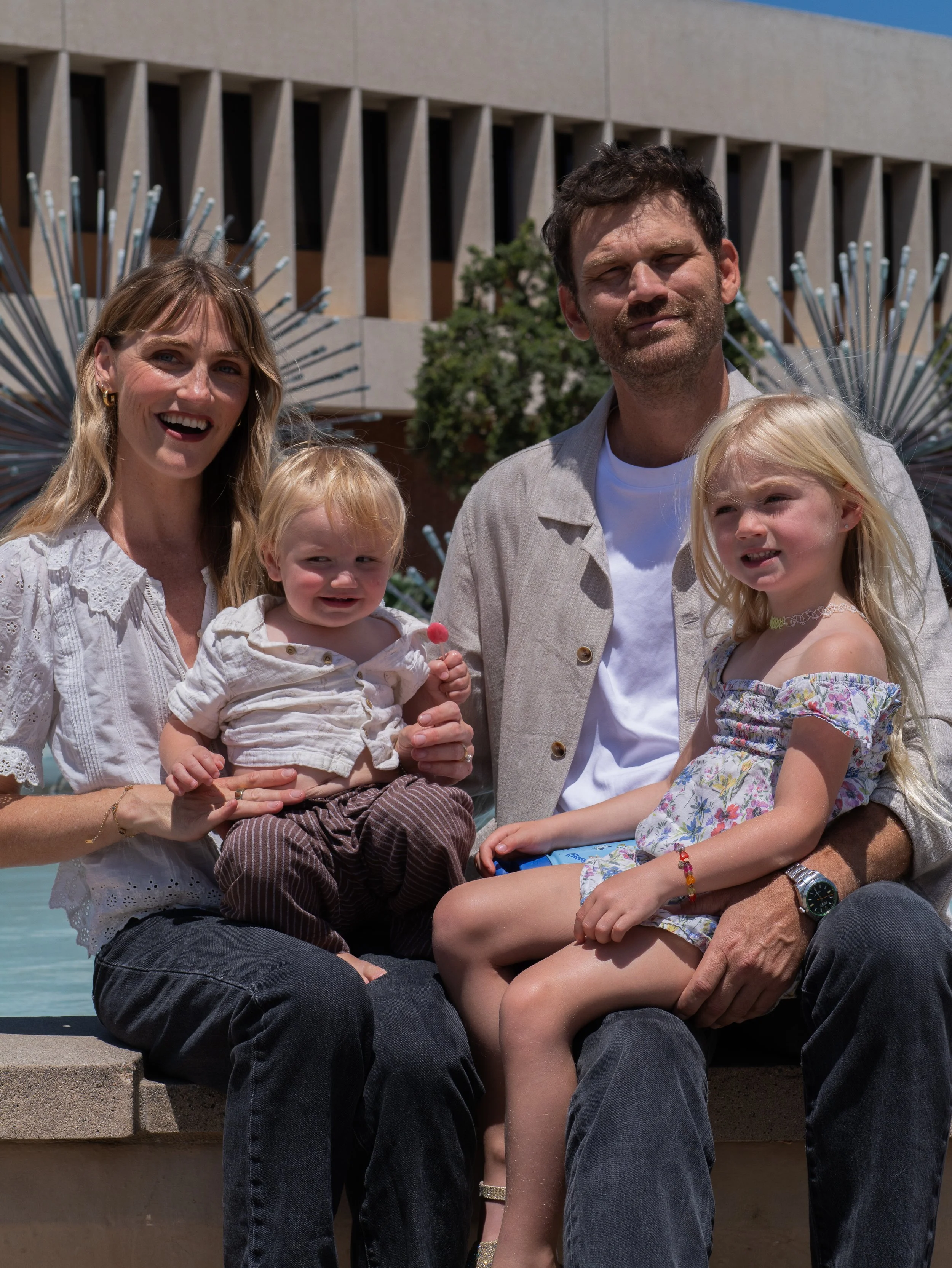 A family of four sitting on a fountain ledge outdoors, with a modern building and water fountain in the background. The woman has long blonde hair, and the man has dark hair and a beard. Both children have blonde hair and are sitting on their parents