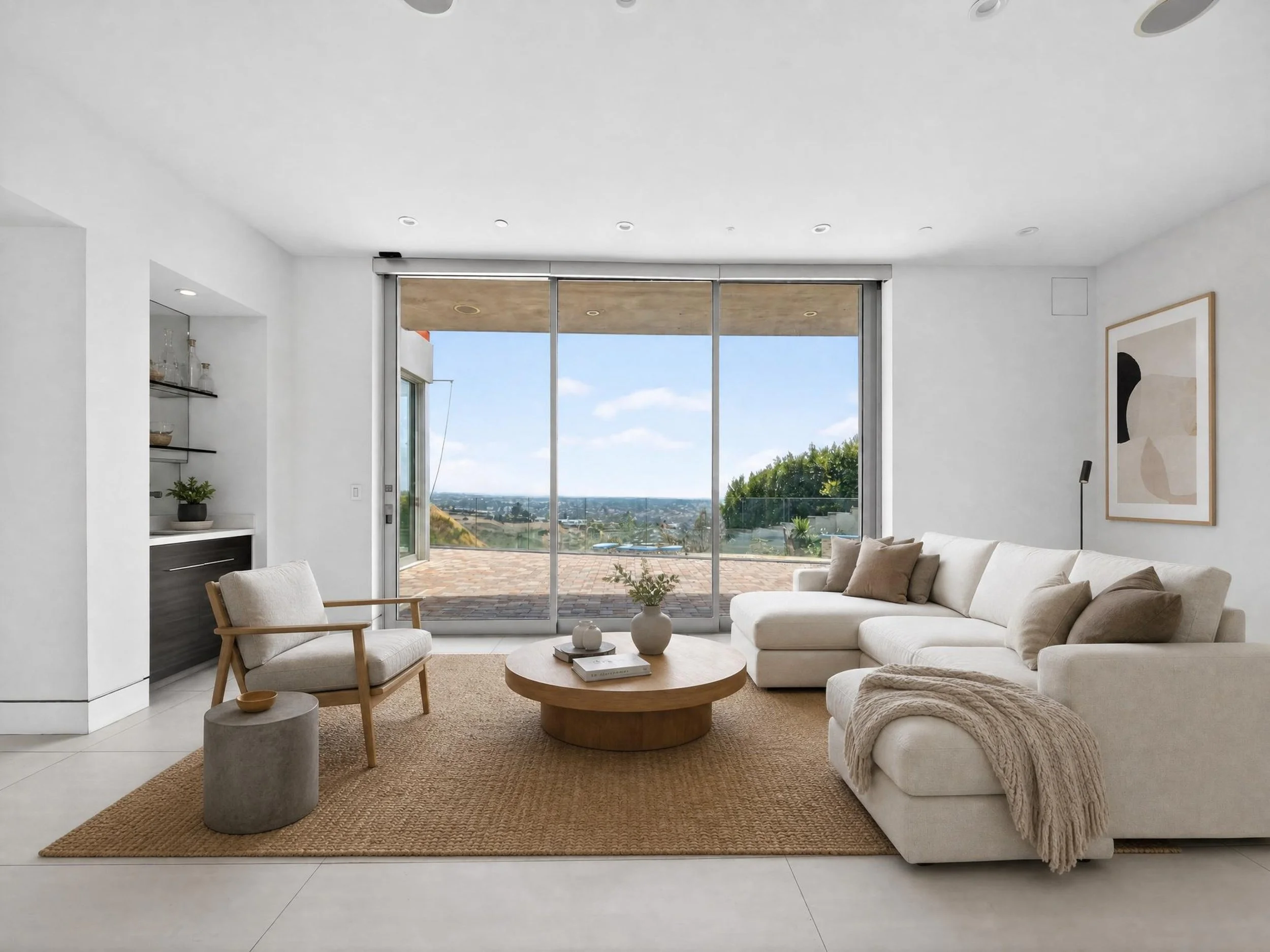 Modern living room with large glass sliding doors showing outdoor patio and landscape, white sectional sofa with beige pillows, wooden coffee table, armchair, framed artwork, and minimal decor.