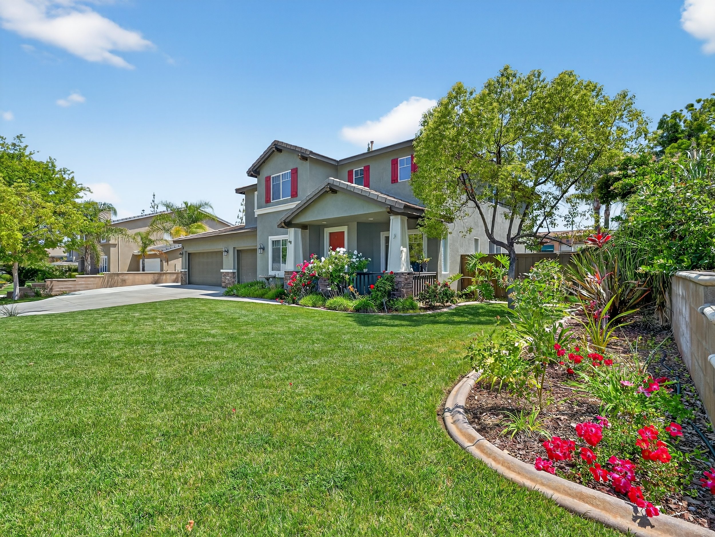 A two-story gray house with red window shutters and a red front door, surrounded by green trees and a well-maintained lawn. A flower bed with pink and red flowers lines the edge of the lawn along a curved stone border.
