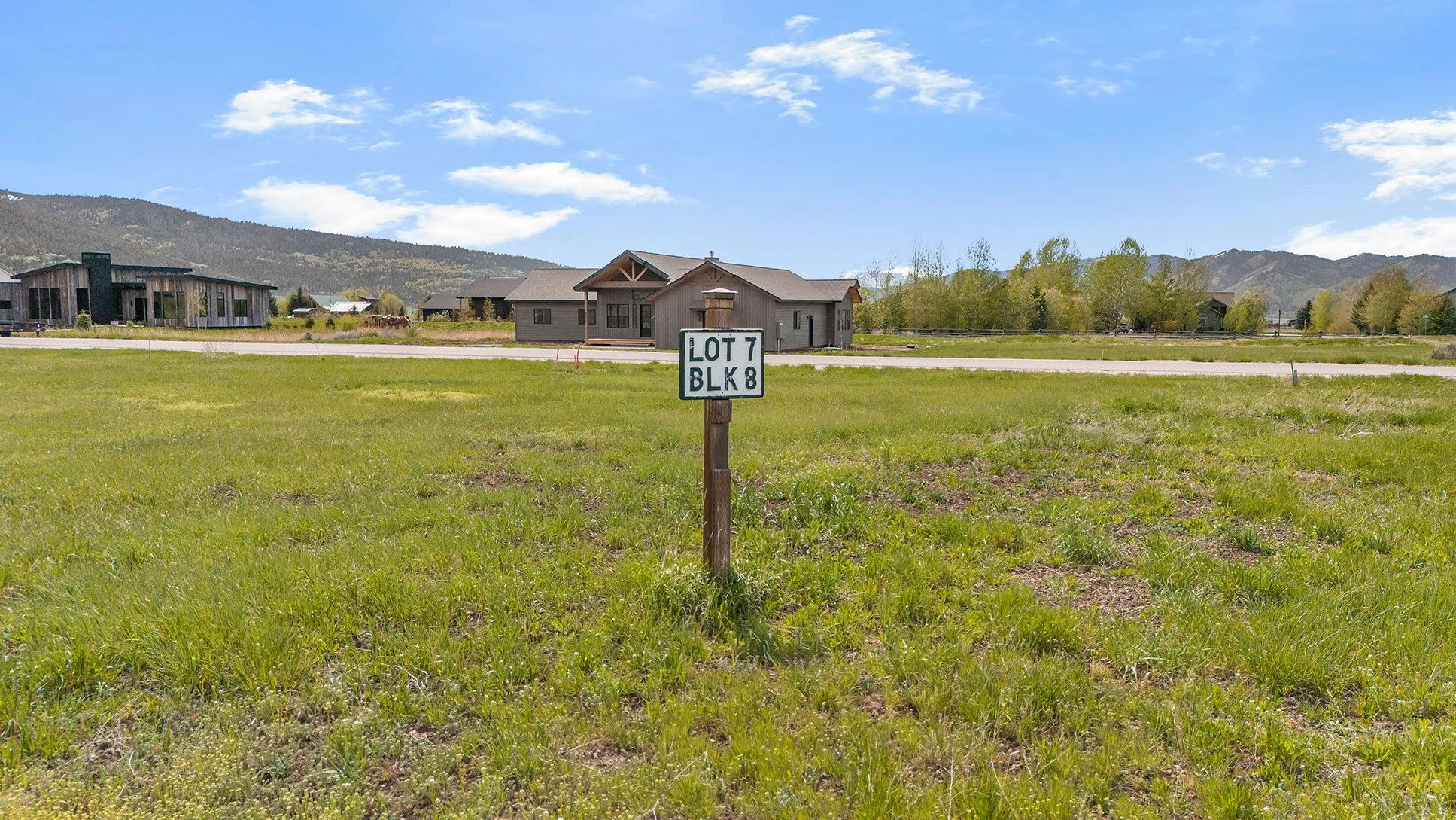 A grassy lot with a street sign reading 'LOT 7 BLK 8' in the foreground. Residential houses with mountains in the background, under a partly cloudy sky.
