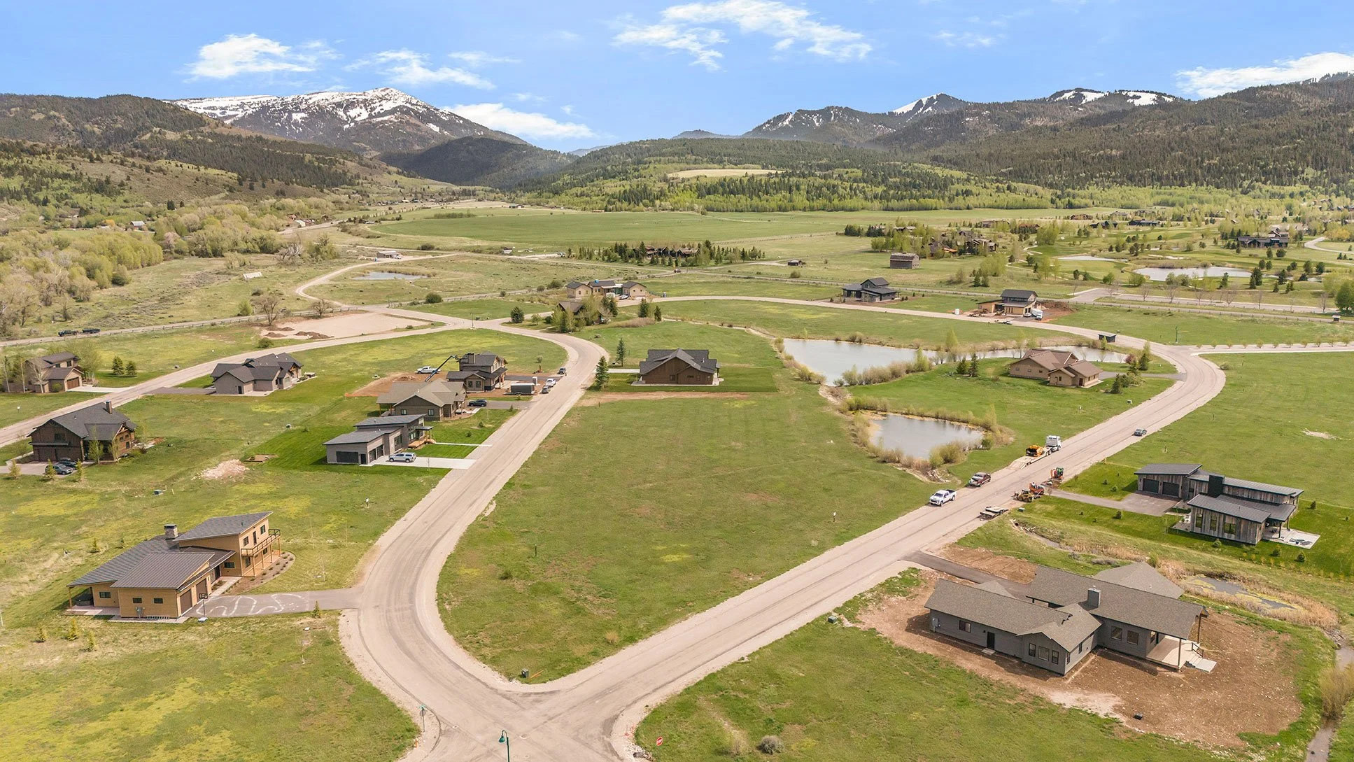 An aerial view of a suburban area with houses, roads, and small ponds, with mountains in the background and a partly cloudy blue sky.