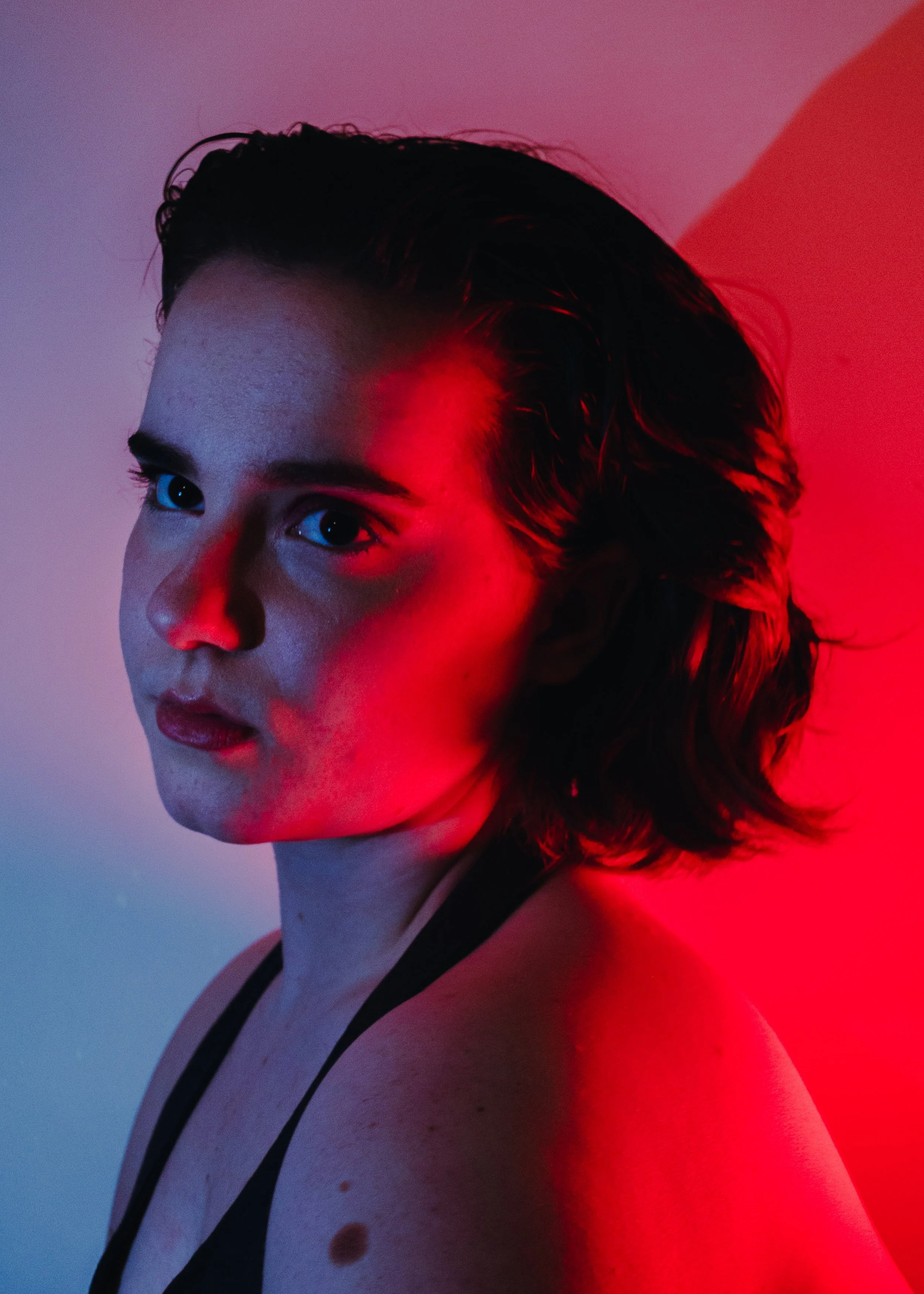 Close-up portrait of a woman with short, dark, wavy hair, looking to her left, illuminated with red and blue lighting, wearing a black top with thin straps.