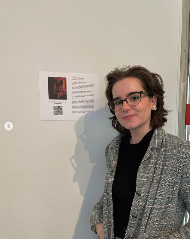 A woman with short brown hair, glasses, and earrings standing next to a wall with a paper posted on it. She is wearing a black shirt and a plaid blazer, smiling slightly.