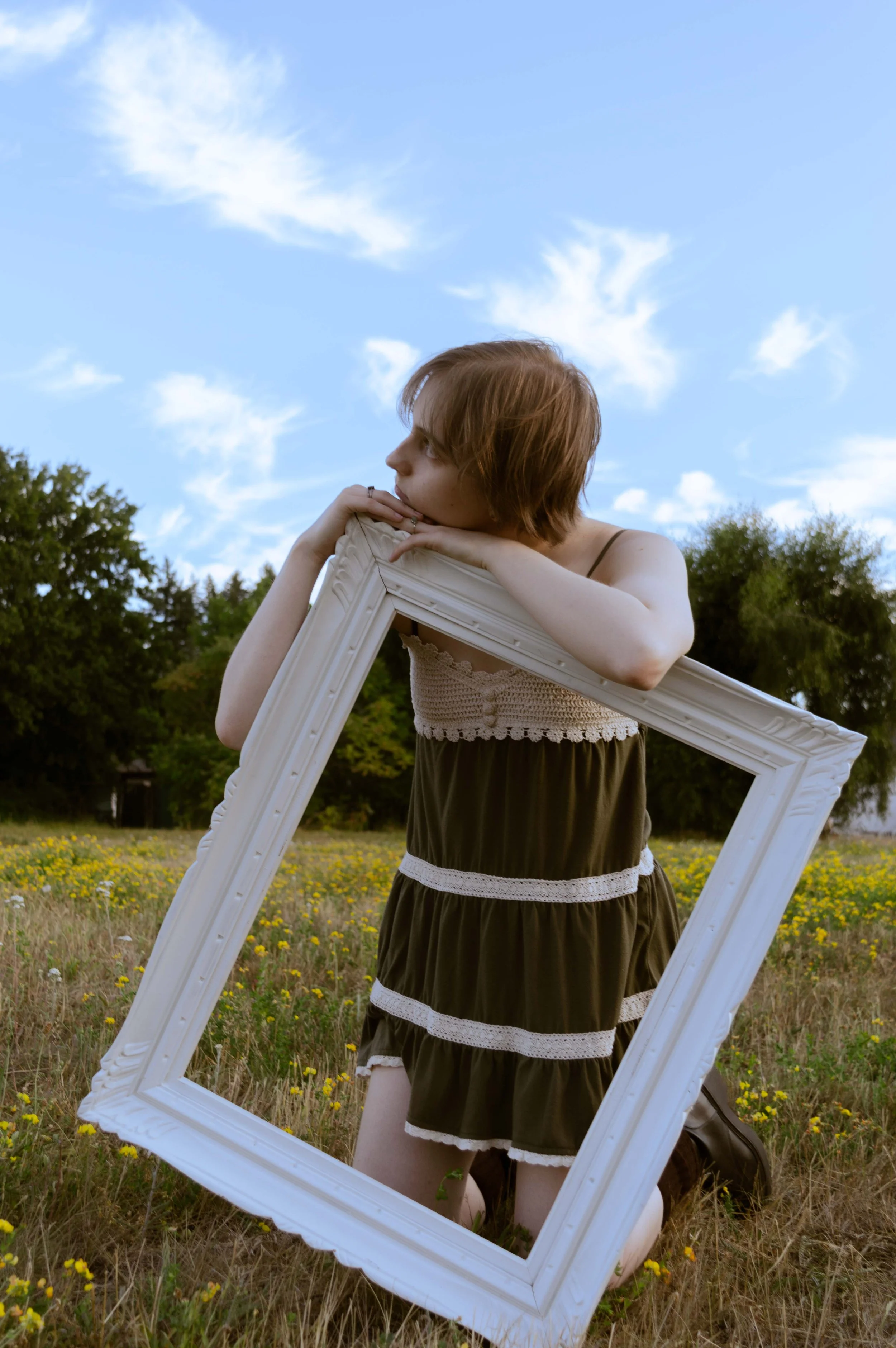 A young woman with short red hair, wearing a strapless dress, is sitting on the grass in a field of yellow flowers while holding a large white picture frame. She is leaning on the frame with her chin resting on her hands, looking to her left. The sce