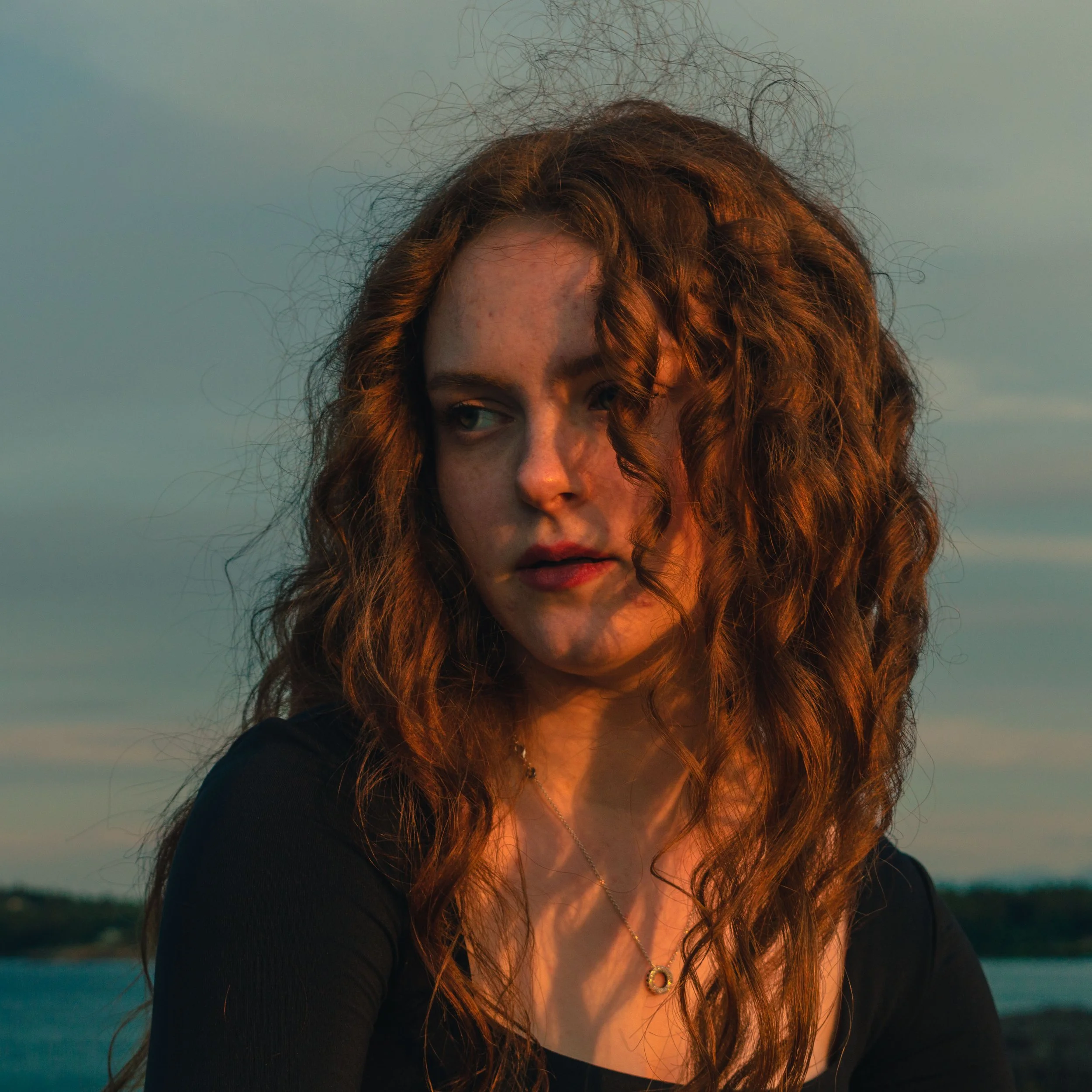 A young woman with long, curly red hair looking to her right, with a sunset or sunrise background.