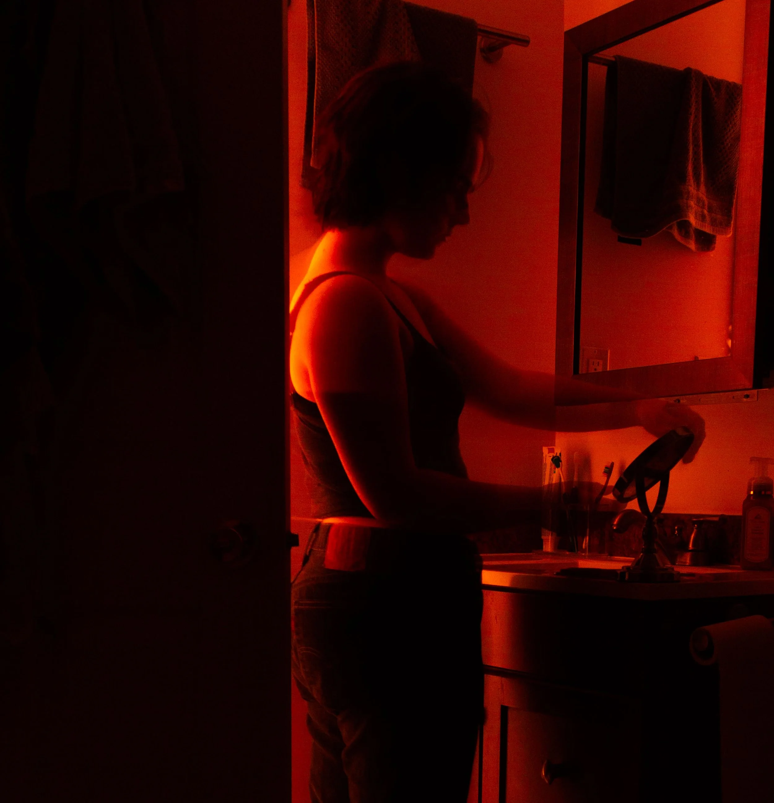 A person with short hair wearing a tank top standing in a room with red lighting, seen in profile, in front of a mirror and a towel hanging on a rack.