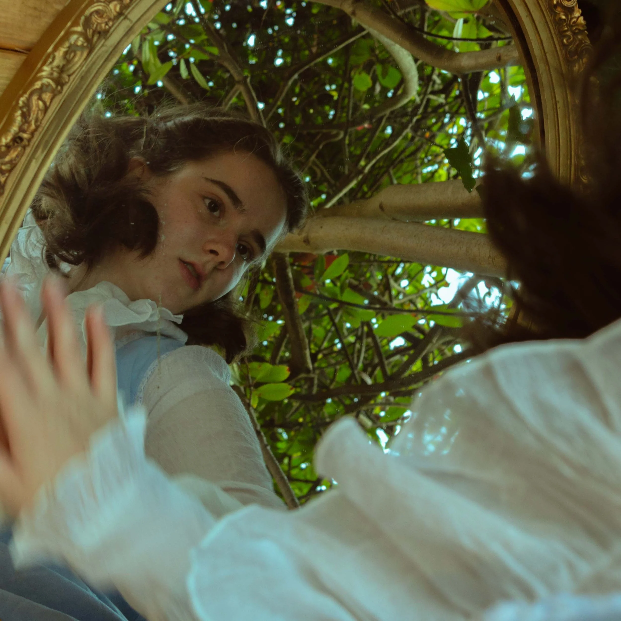 A woman with brown hair wearing a white top taking a selfie in front of a mirror reflecting nearby green foliage and tree branches.