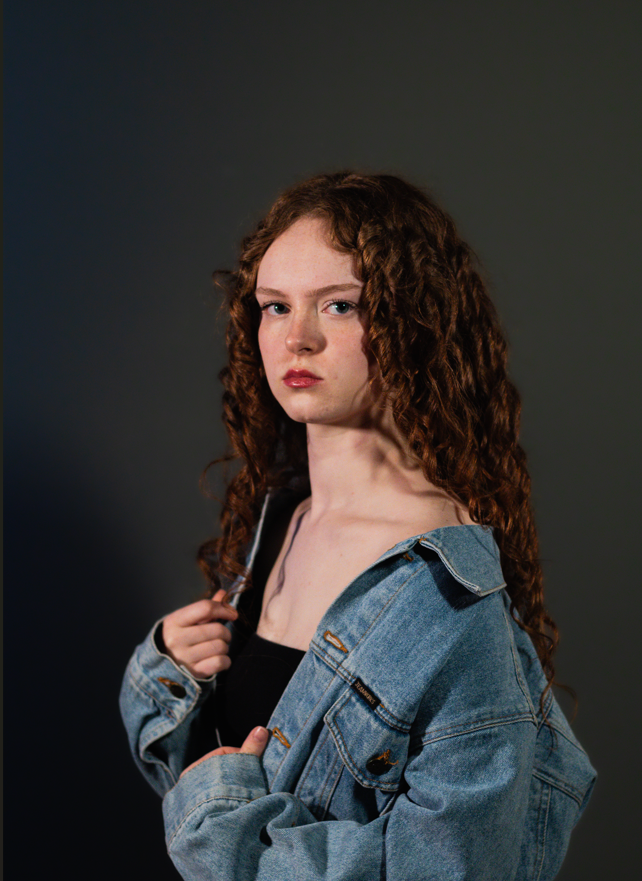 Young woman with curly red hair wearing a denim jacket and black top, holding a small knife, against a dark background.
