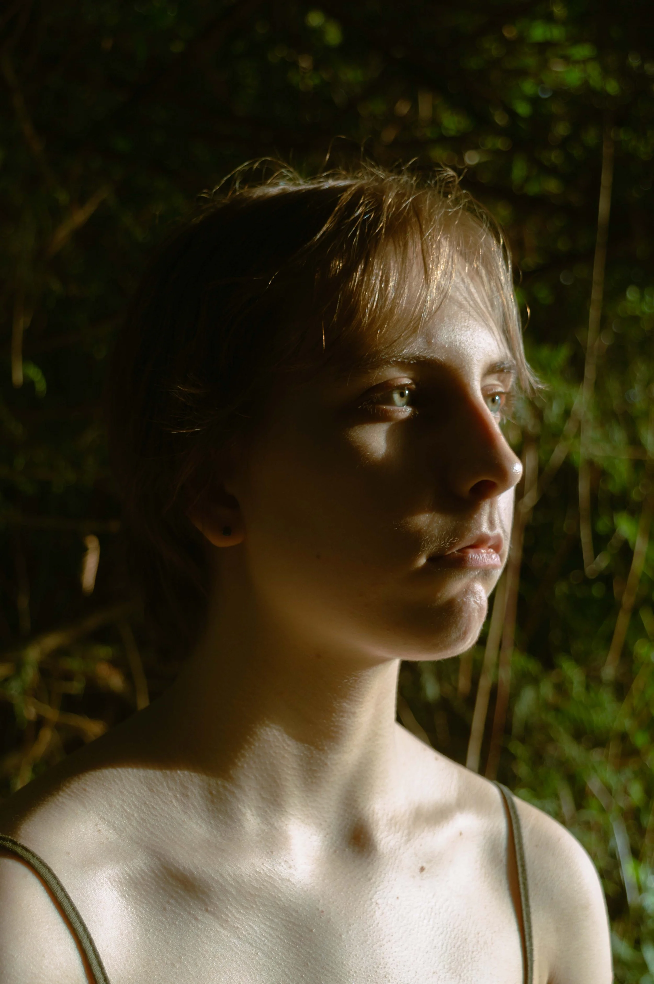 Close-up of a young woman with blond hair, blue eyes, and fair skin, looking to her right against a background of green foliage.