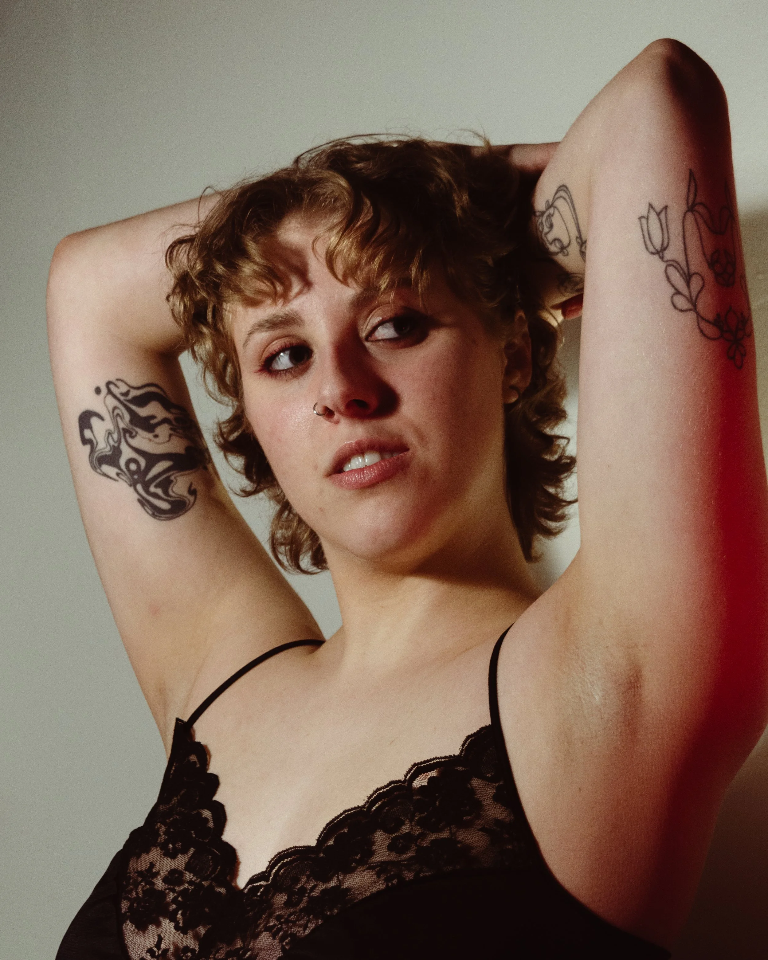 Close-up of a woman with short, curly red hair and tattoos on her arms, wearing black lace lingerie with a floral pattern, posing with her arms raised behind her head against a neutral background.
