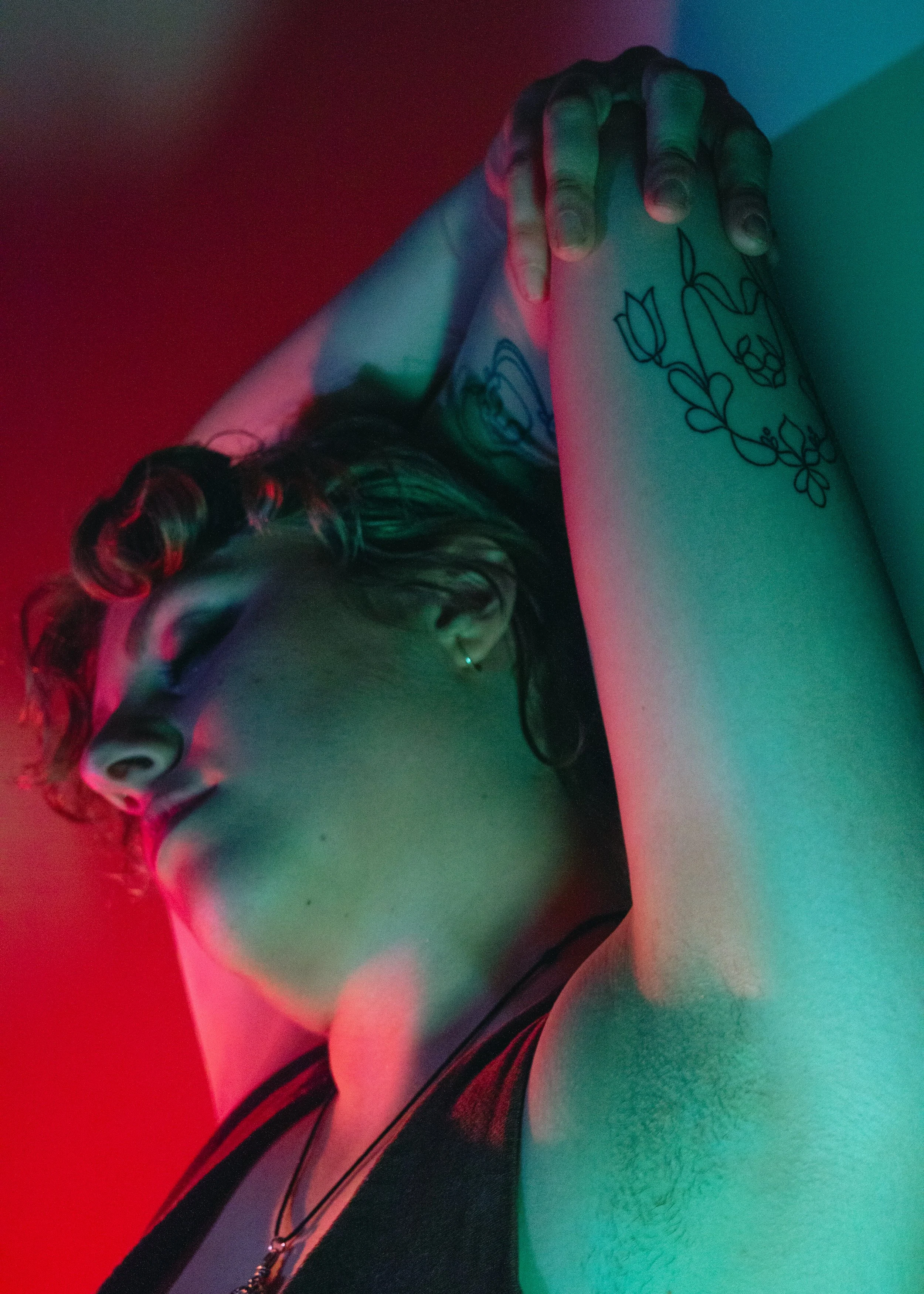 A person with dark curly hair, a nose ring, and a necklace, standing against a background with red and teal lighting.