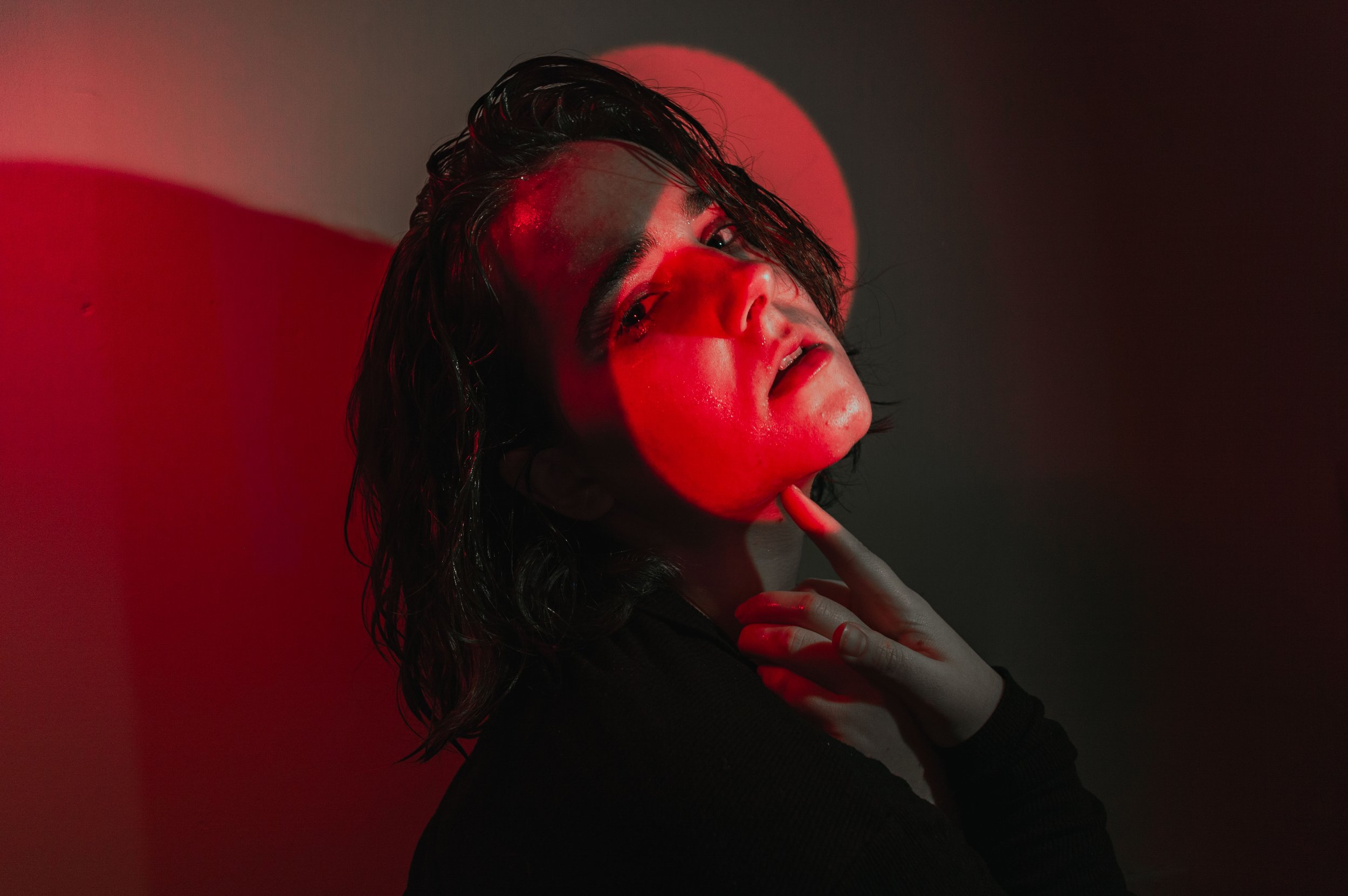 Portrait of a woman with wet dark hair, holding her chin with one hand, illuminated by red and black lighting.