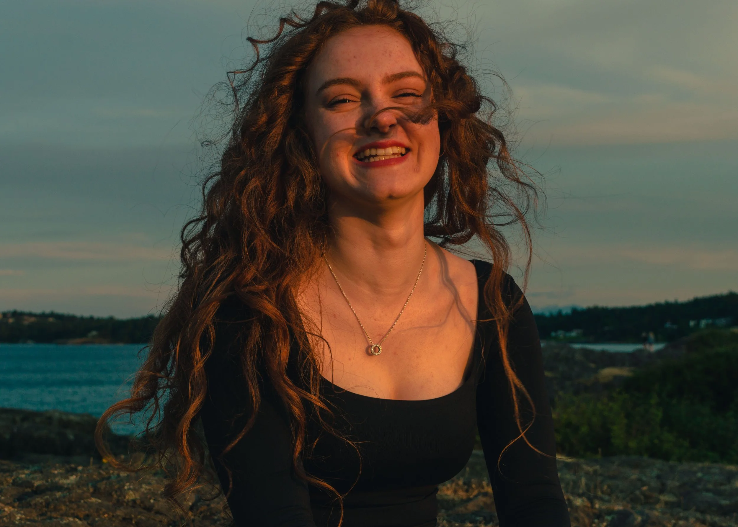 A young woman with curly red hair and freckles is smiling outdoors at sunset, wearing a black top and a silver necklace with a circular pendant, with a body of water and a hilly landscape in the background.