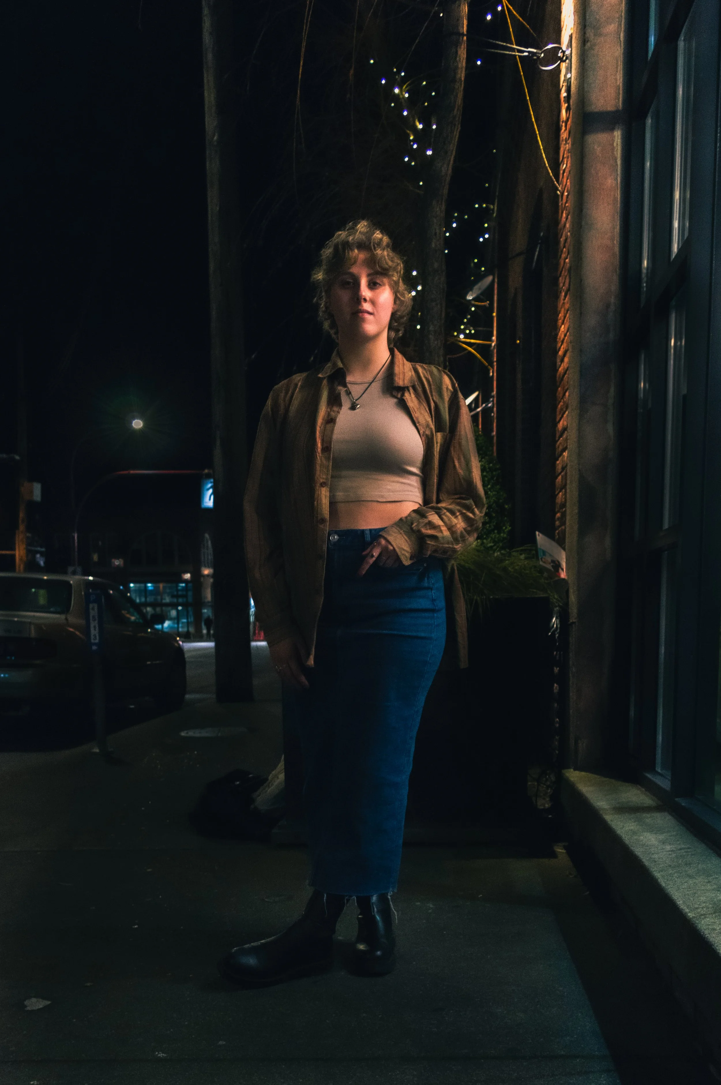 A young woman standing on a city sidewalk at night, dressed in a brown plaid shirt over a white crop top and a long blue skirt, wearing black boots and a necklace, with streetlights, parked cars, and illuminated buildings in the background.