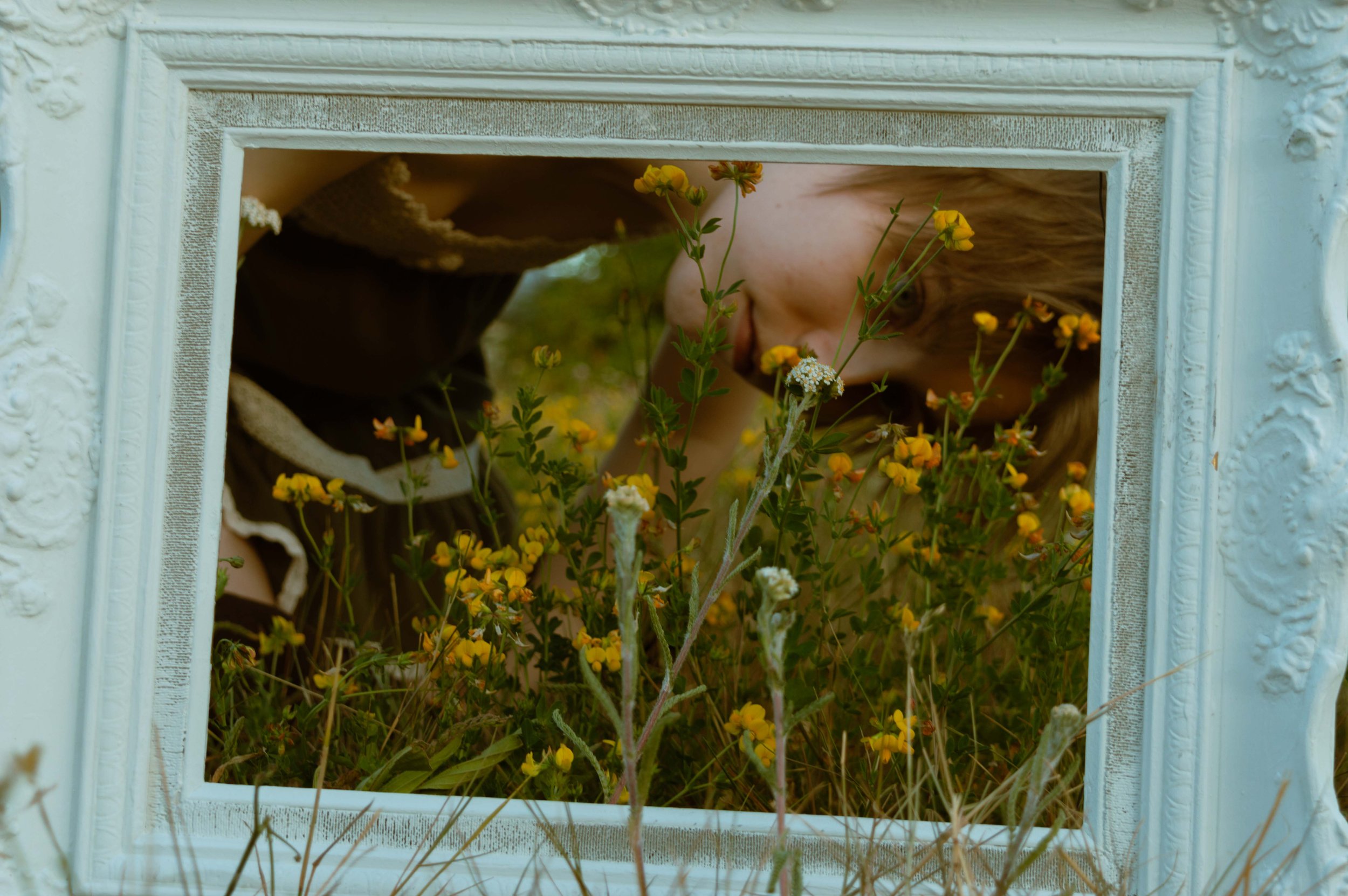 Person crouching behind a white ornate mirror frame with yellow and white flowers growing through the opening.