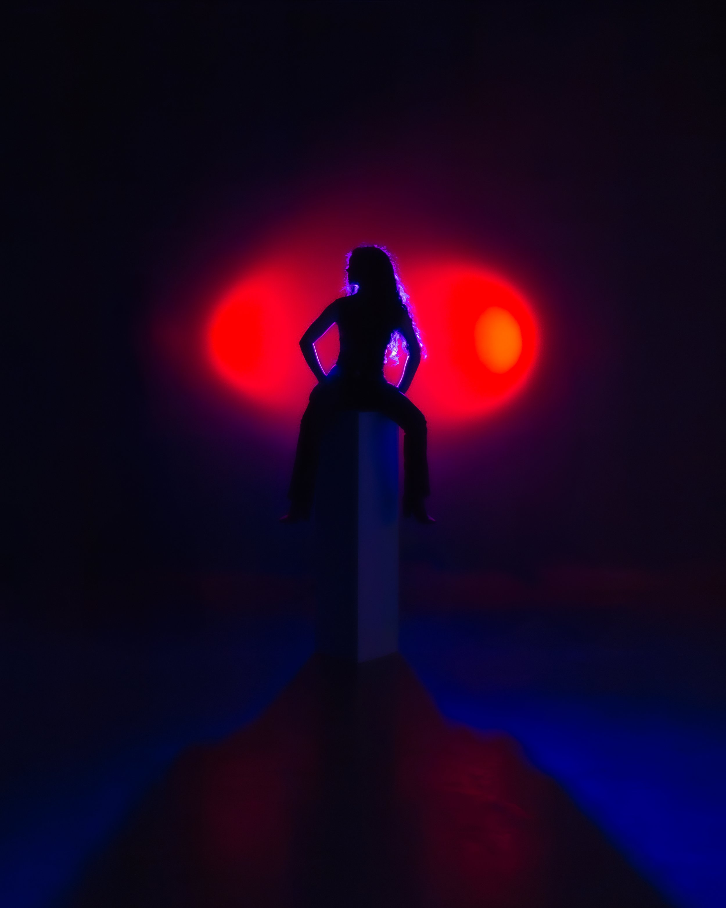 Silhouette of a woman with curly hair sitting on a stool against a dark background illuminated by red and blue spotlight effects.