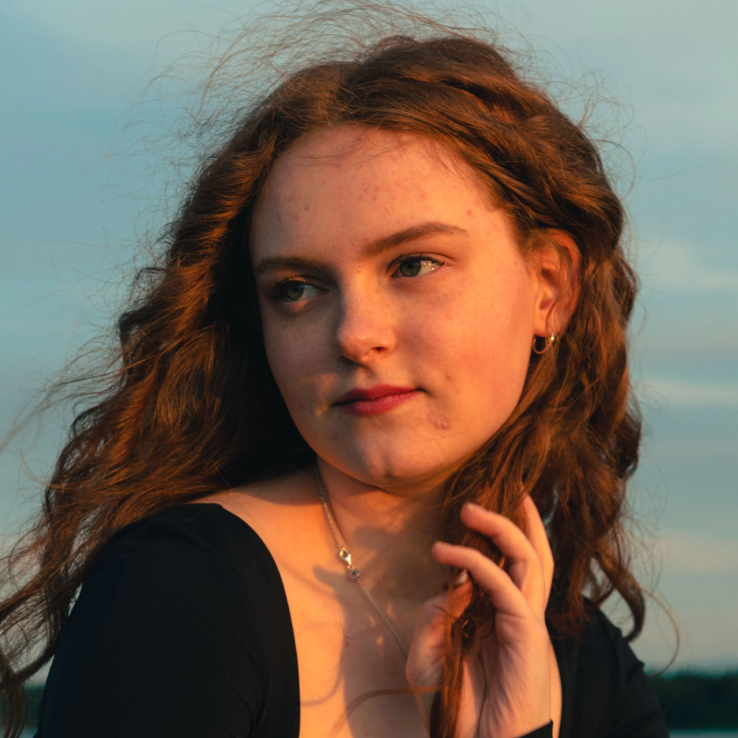 A woman with curly auburn hair and blue eyes, wearing a black top and a silver necklace, stands outdoors during sunset with a green landscape and sky in the background.