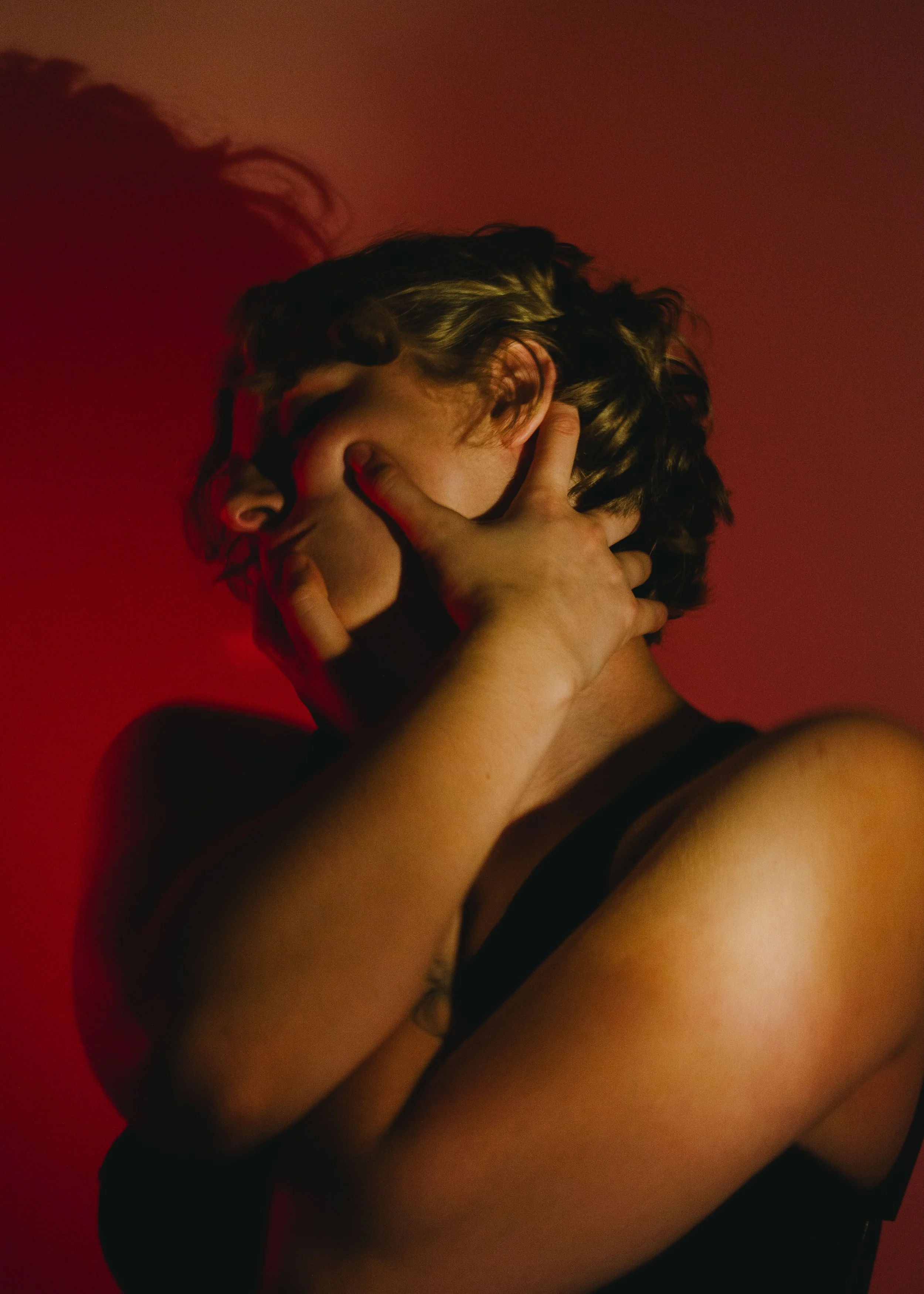 A person with short curly hair, wearing a black sleeveless top, is covering their face with their hand. The background is red.