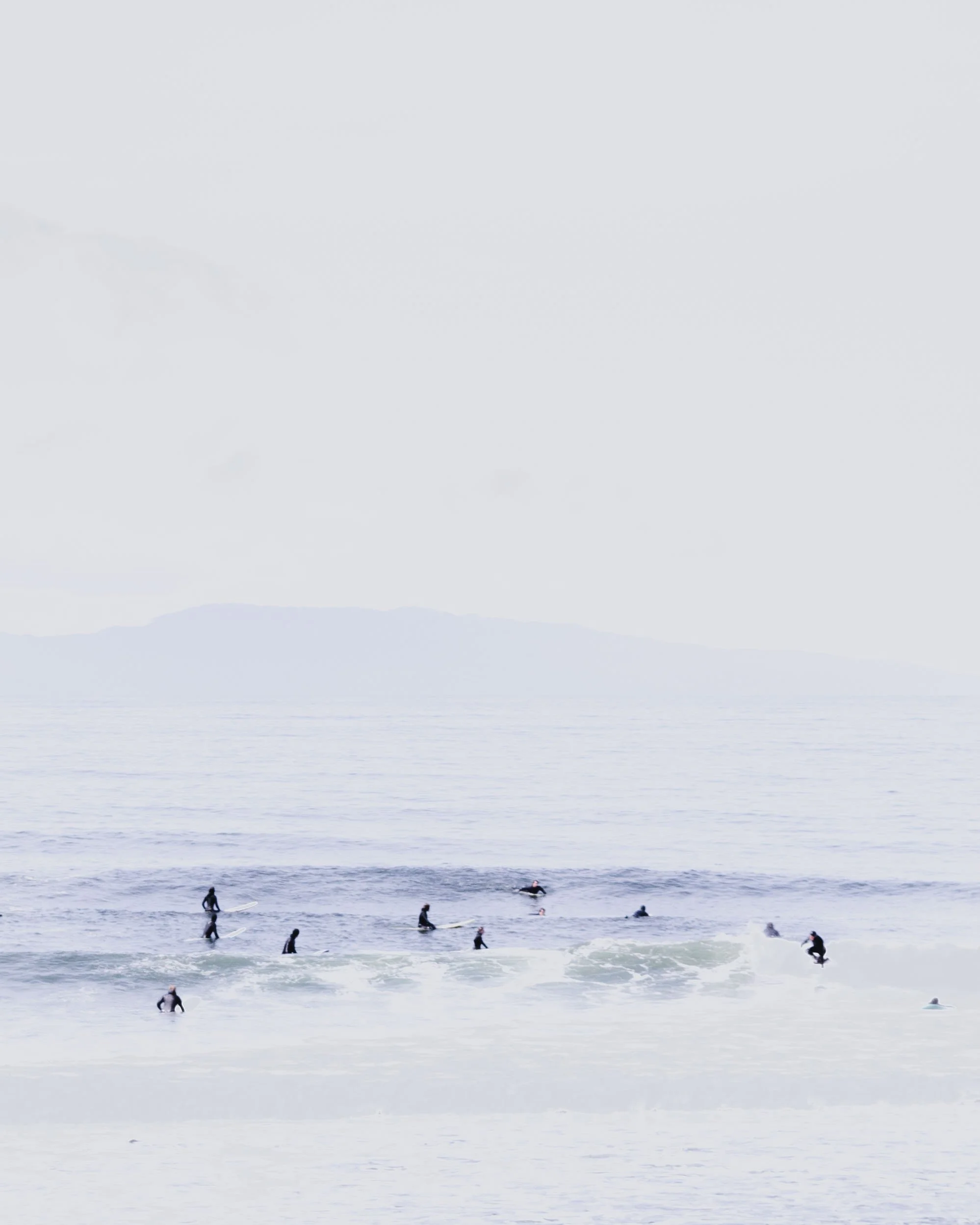 Surfers in wetsuits riding and waiting for waves at the beach under an overcast sky.