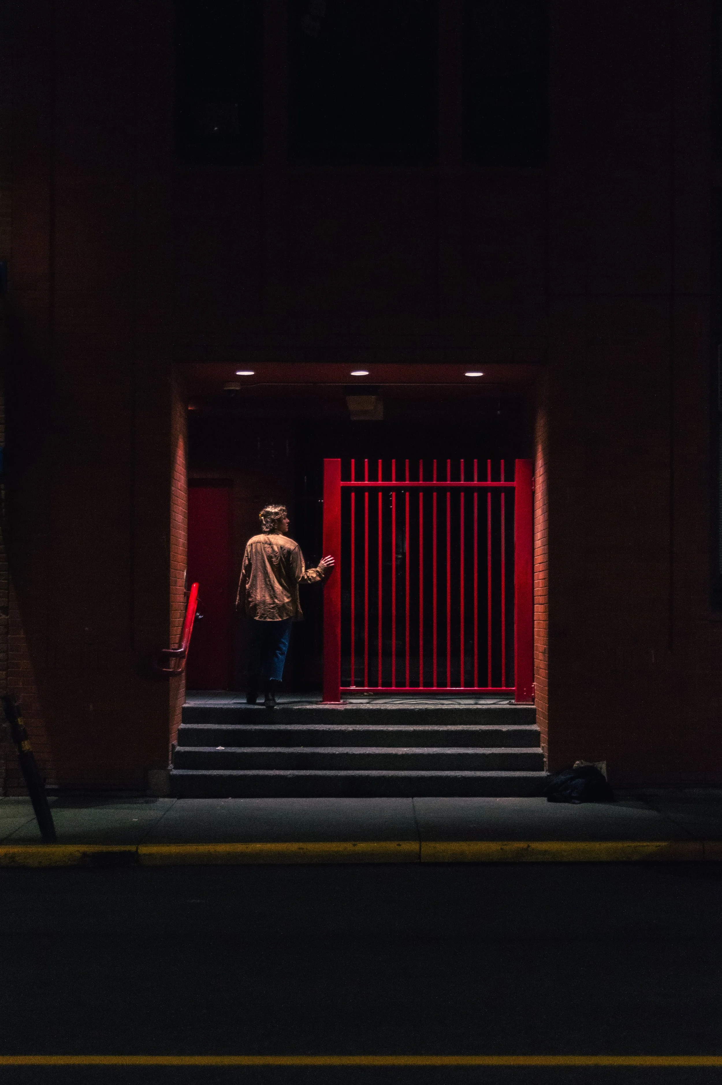 A person with curly hair and a brown jacket standing on steps in front of a red gate at night, with dark walls on either side and a black window above.