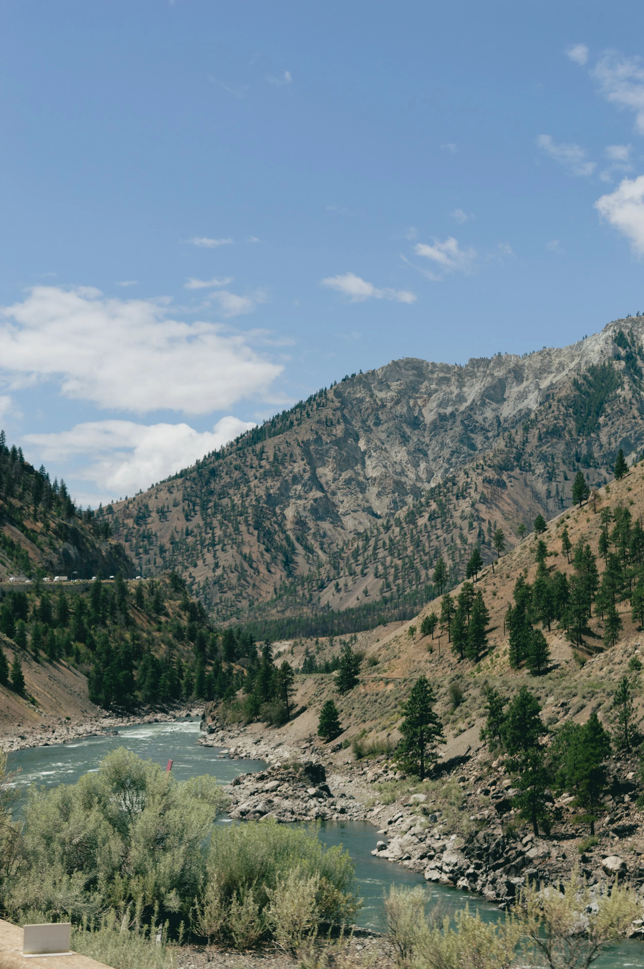 A mountain landscape with a blue sky, sparse clouds, and a river flowing through a valley surrounded by rocky hills and scattered trees.
