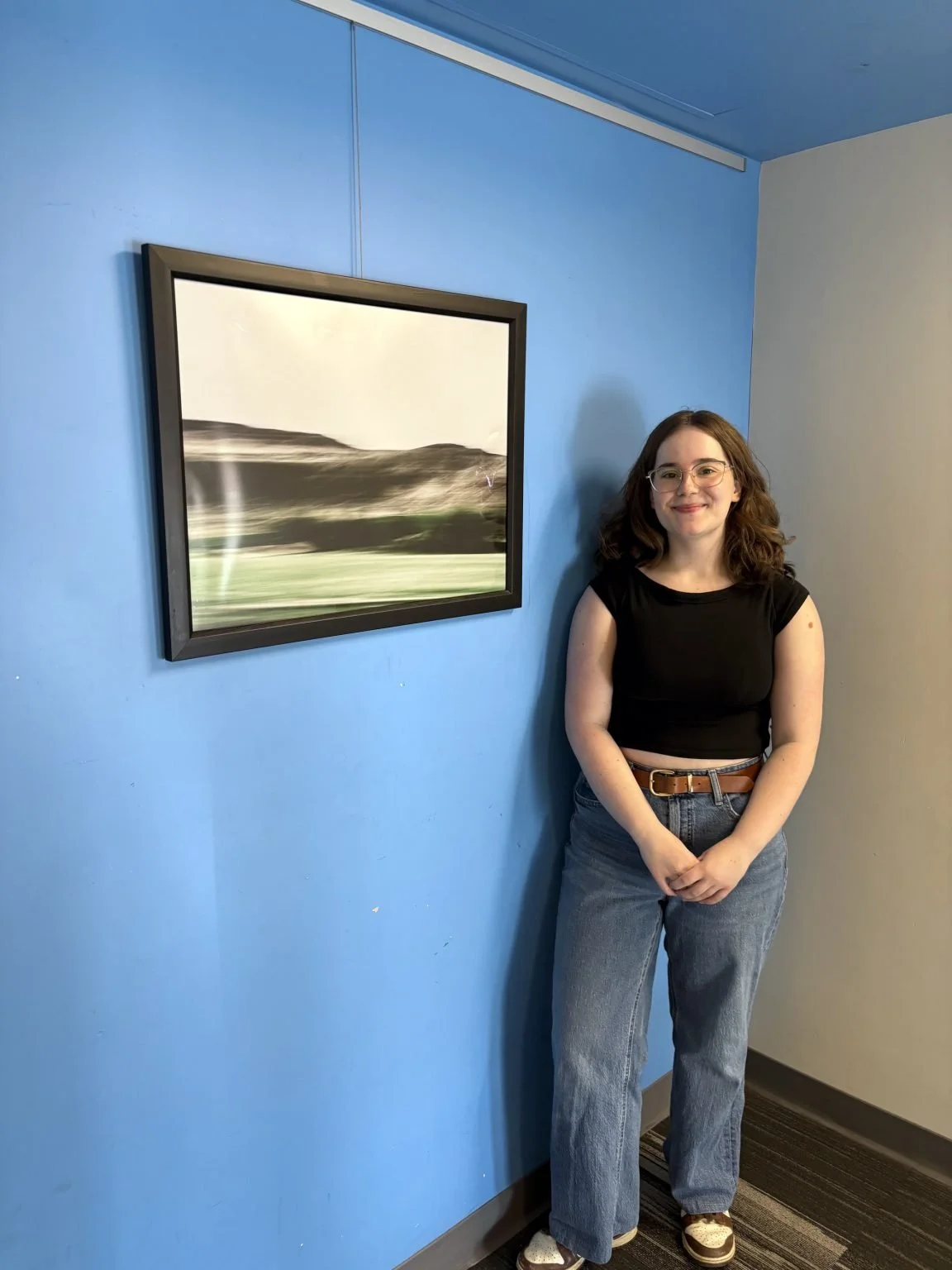 A young woman in casual clothing standing next to a framed landscape photograph on a blue wall in an indoor setting.