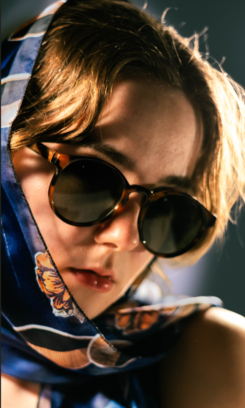 Close-up of a person wearing sunglasses and a blue patterned headscarf, with sunlight highlighting their hair and face.