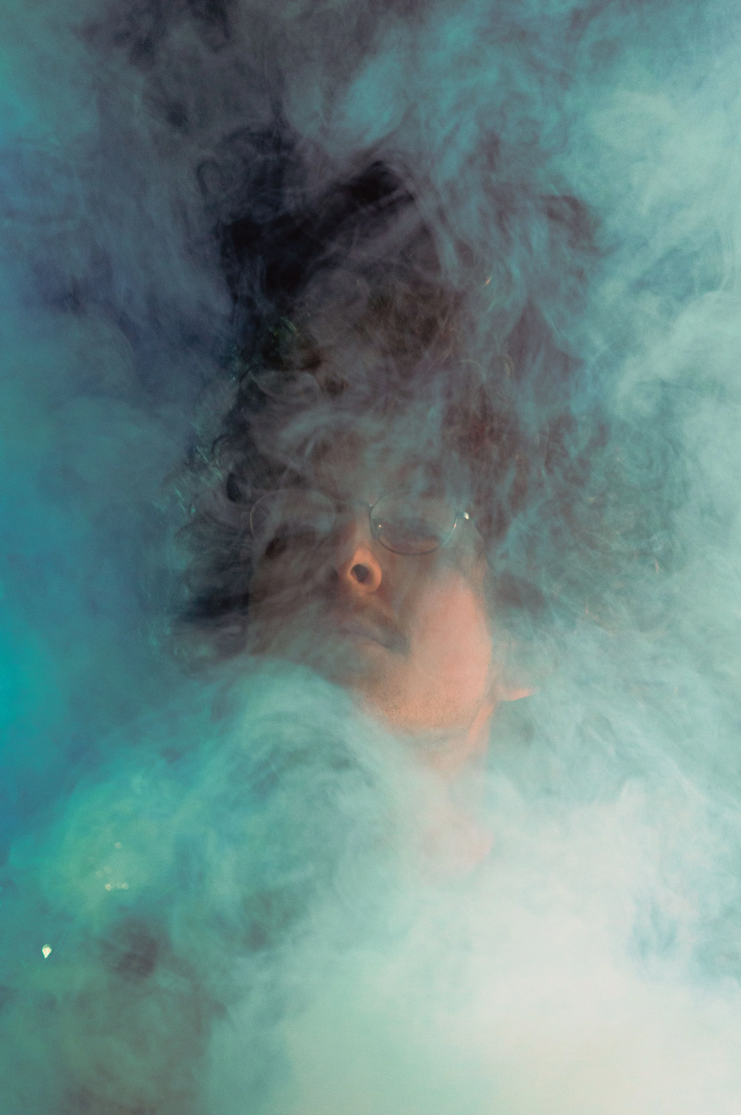 A person's face partially submerged in water with swirling smoke or ink around, creating a mysterious and abstract visual effect.