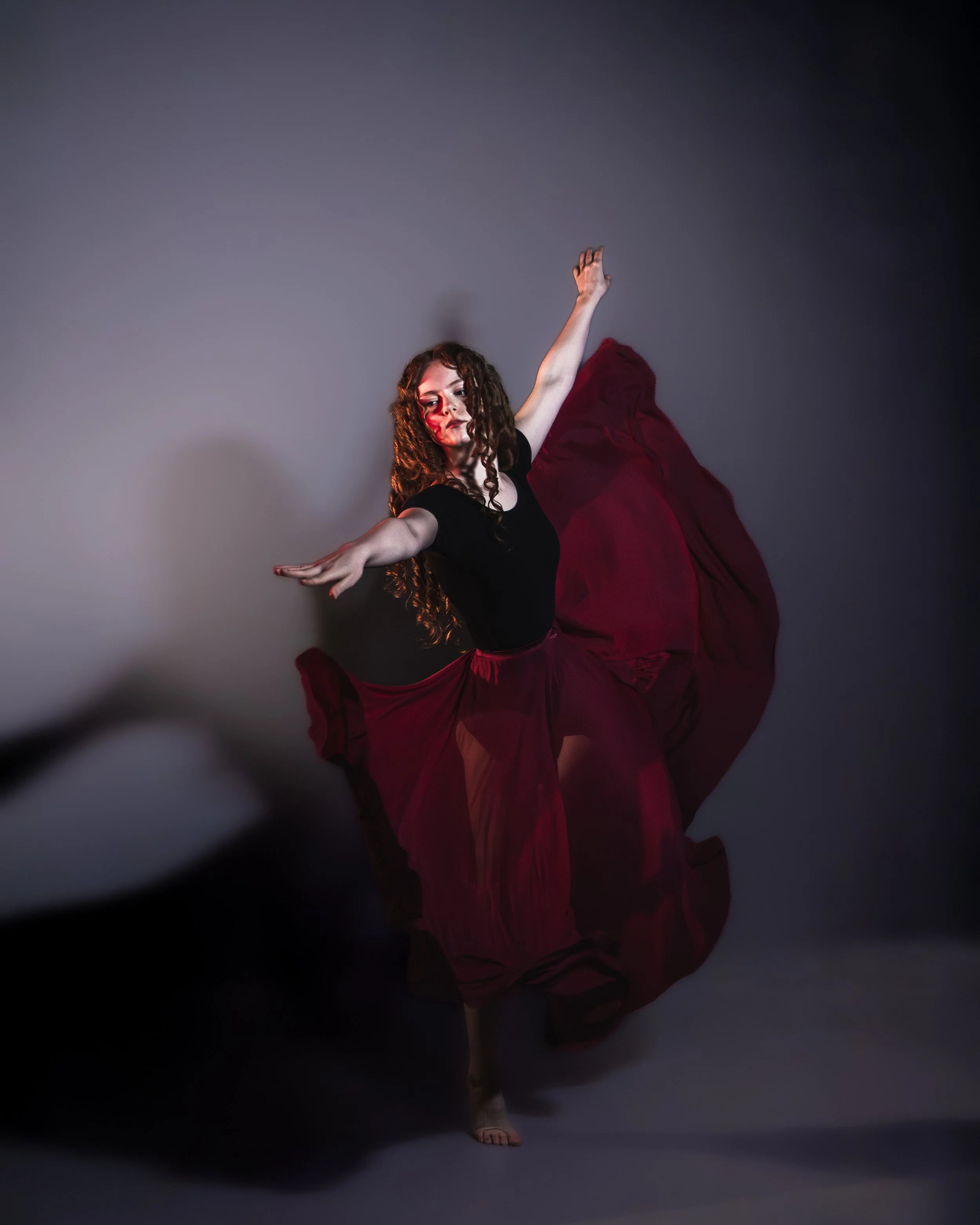 A dancer with curly hair wearing a black top and a long burgundy skirt, balancing on one foot with arms extended, against a gray background.