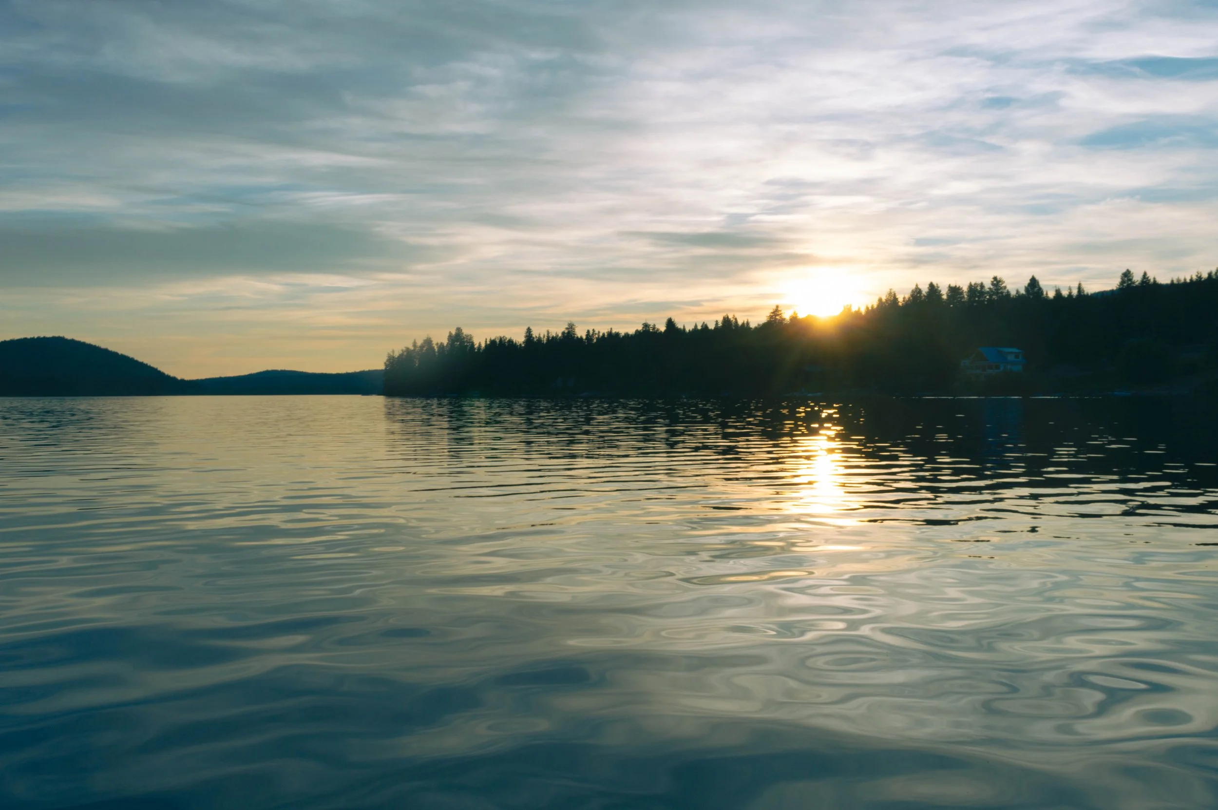 Sunset over a lake with trees on the shoreline and a house visible in the distance.