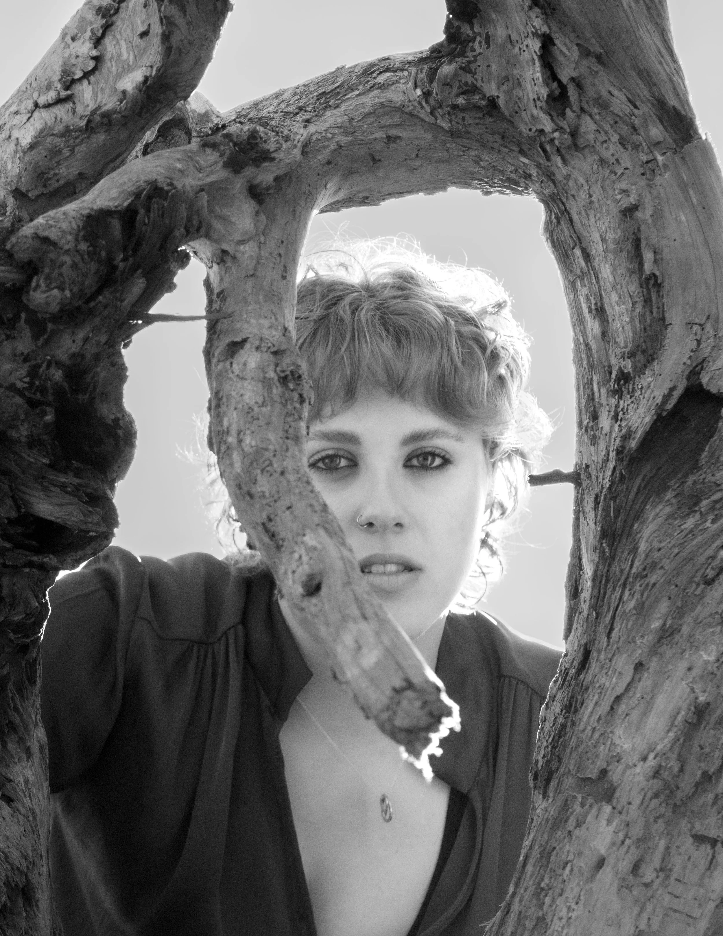 Close-up black and white photo of a person's face partially obscured by twisted tree branches framing the image, with visible eyes, nose, and eyebrows.