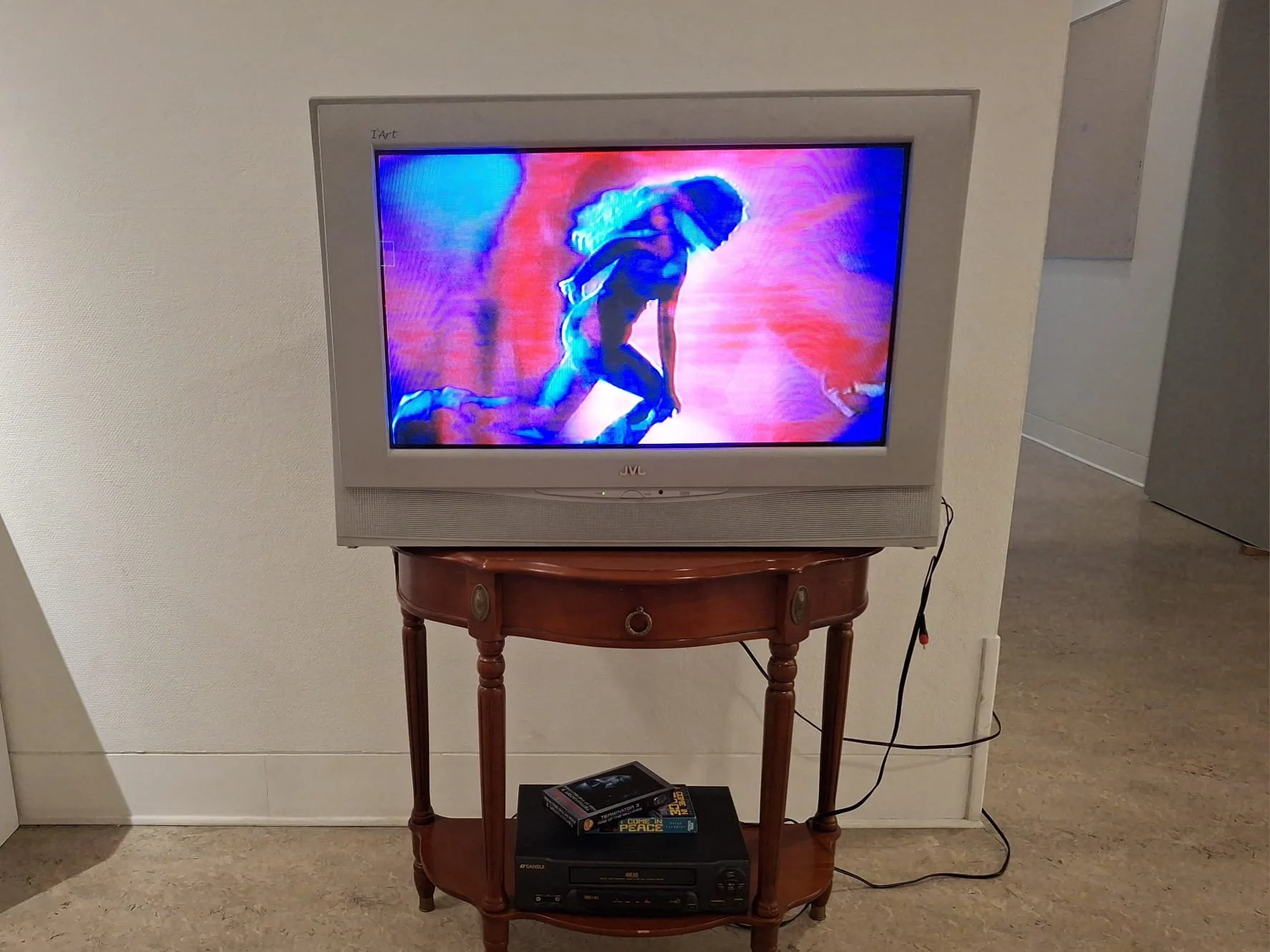 A TV displaying a colorful animated or video game scene, placed on a wooden stand with books and electronic equipment underneath, in a room with beige walls and carpeted floor.
