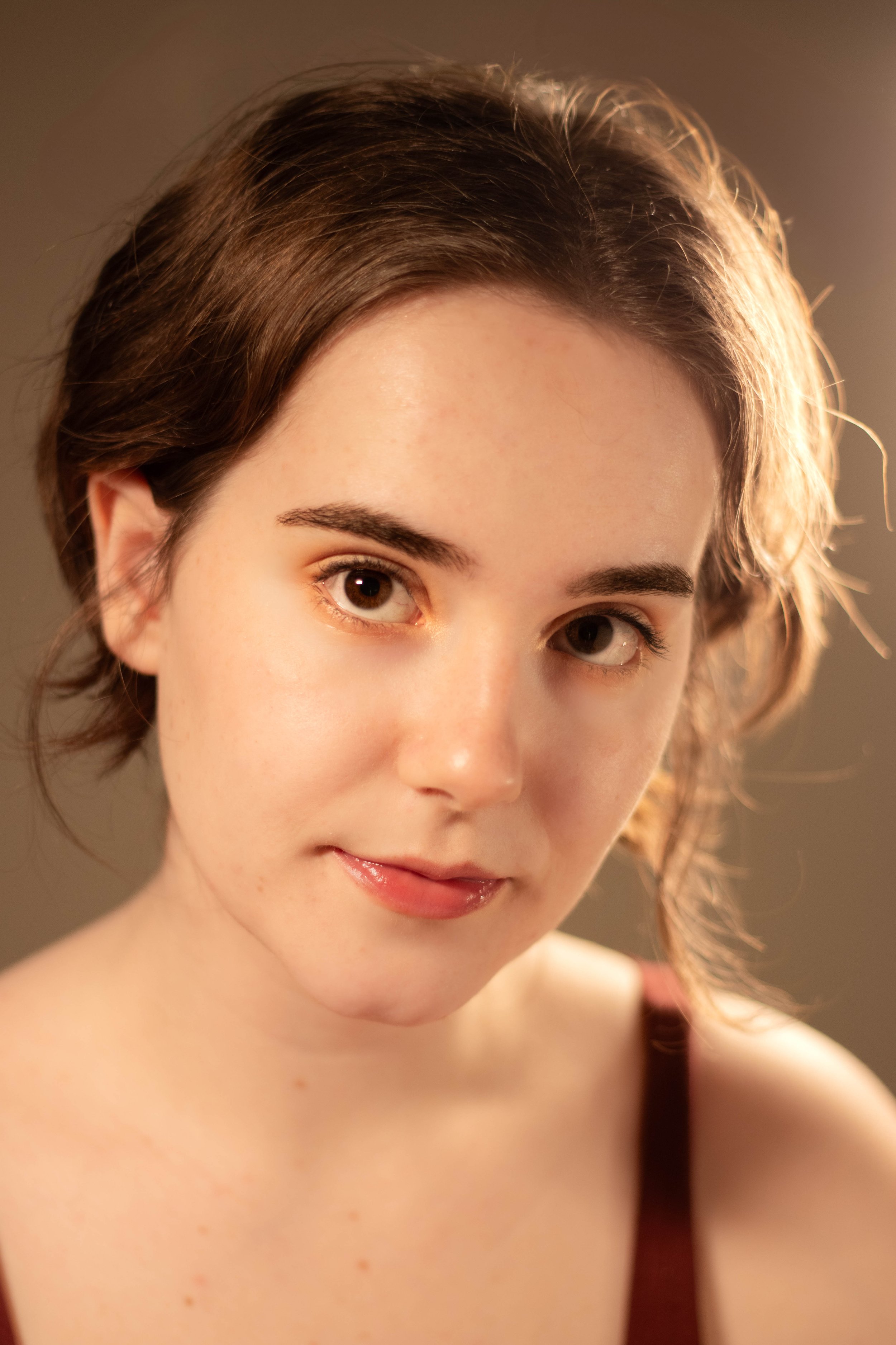Close-up of a young woman's face with brown hair, brown eyes, and a slight smile, wearing a dark-colored top, with soft lighting highlighting her features.