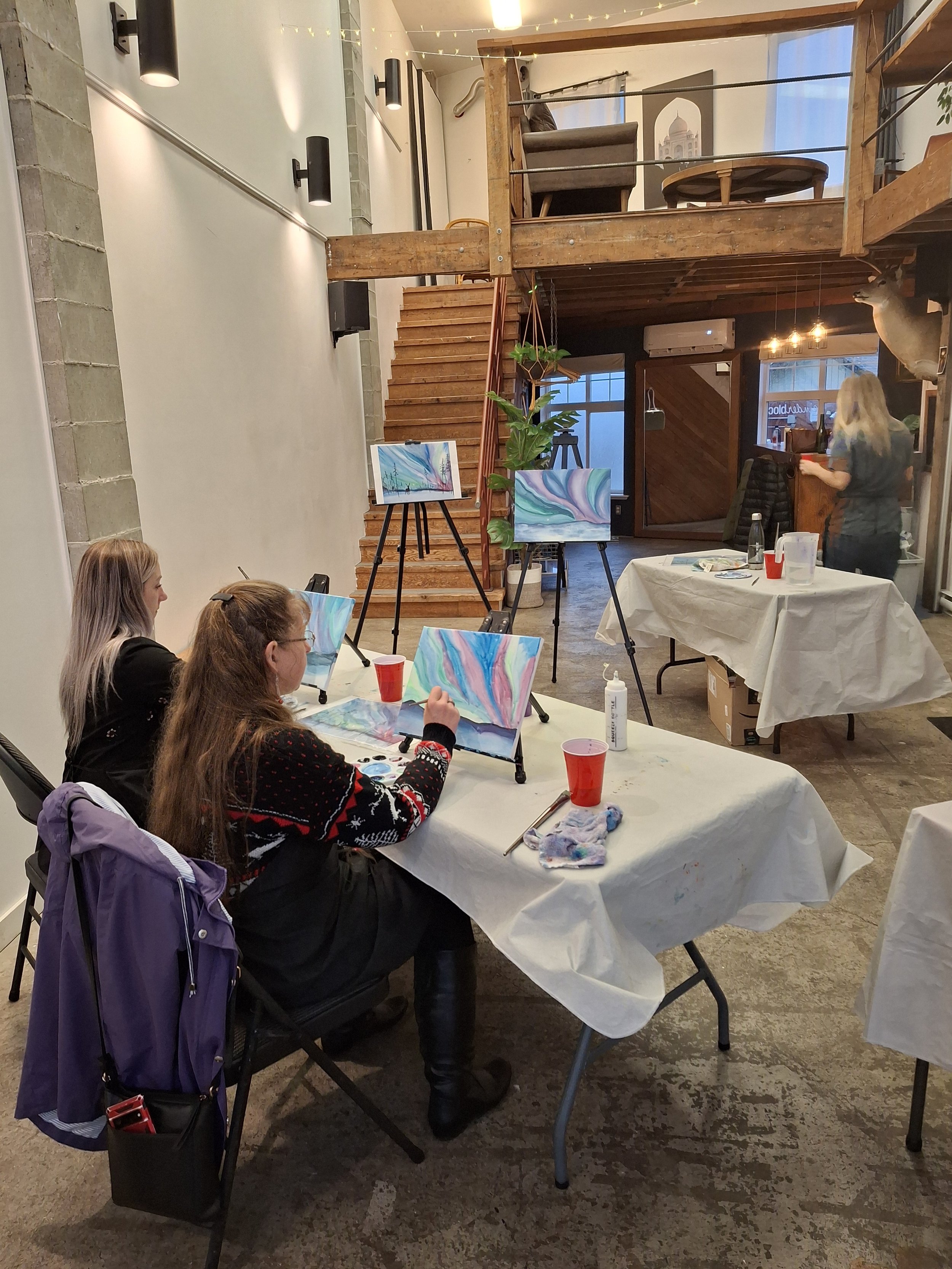 A group of women participating in a watercolor painting class in an industrial-style studio with high ceilings, exposed concrete and wooden accents, some paintings on easels, and a woman standing at the back holding a red cup.