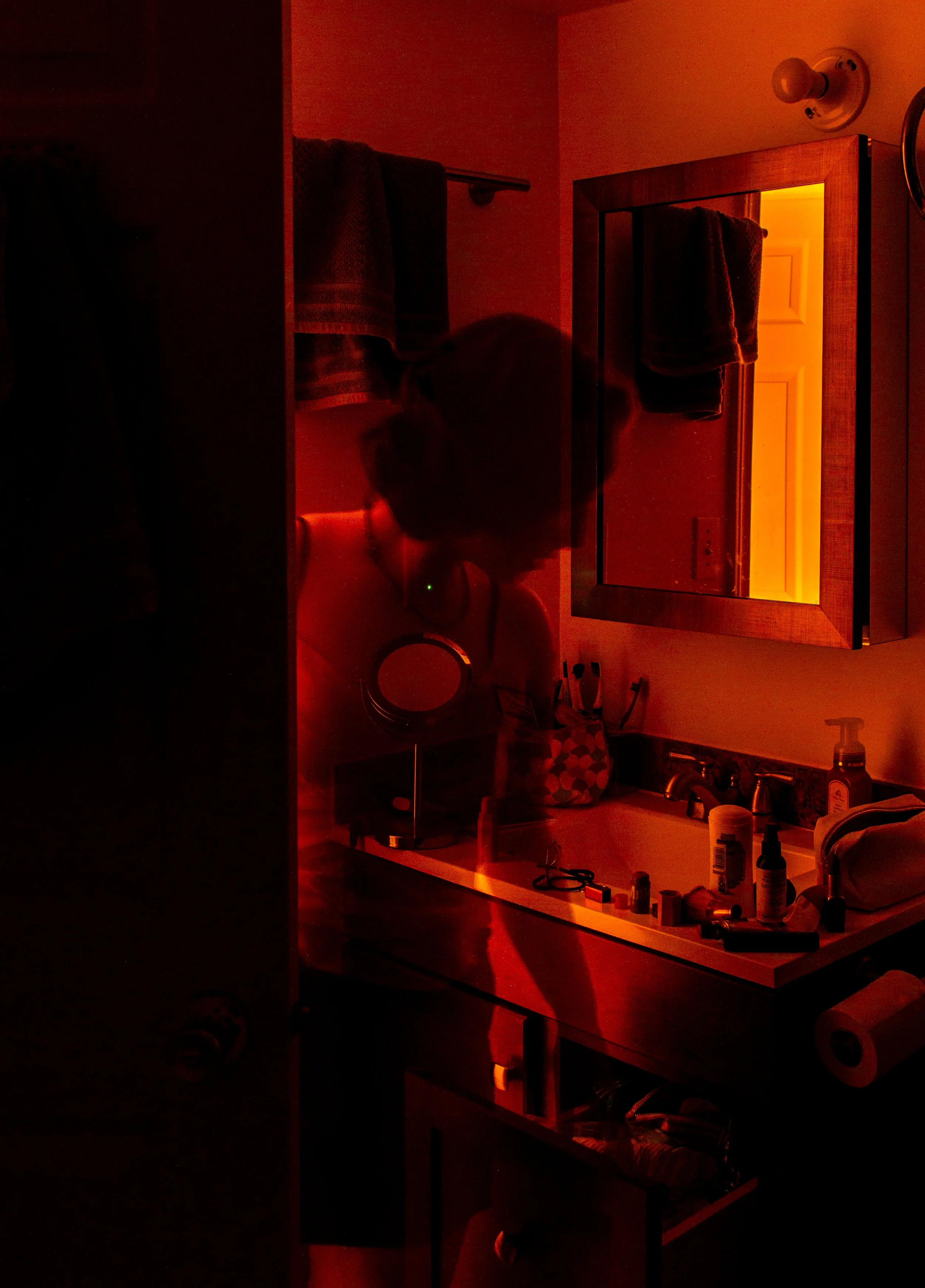 A bathroom mirror and sink area illuminated with red light, with toiletries and small objects on the counter, a towel hanging on the wall.