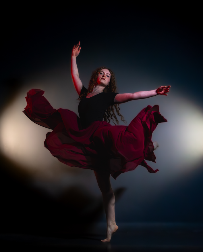 A female dancer performing a ballet move, wearing a black top and a flowing red skirt, against a dark background.
