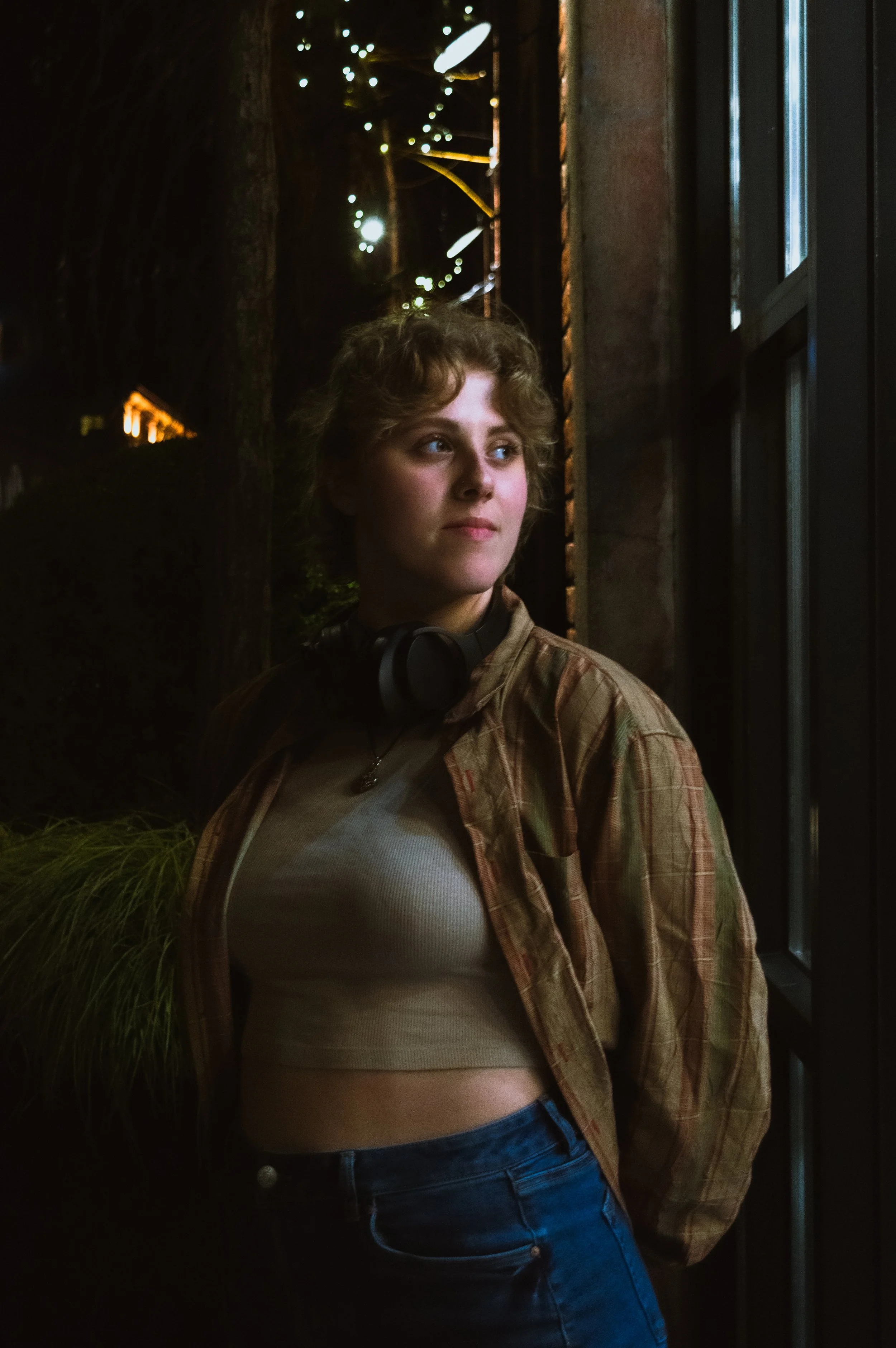 A young woman with short, curly hair looking out of a window at night. She is wearing a plaid shirt over a crop top and has headphones around her neck. The background is dark with some city lights visible.