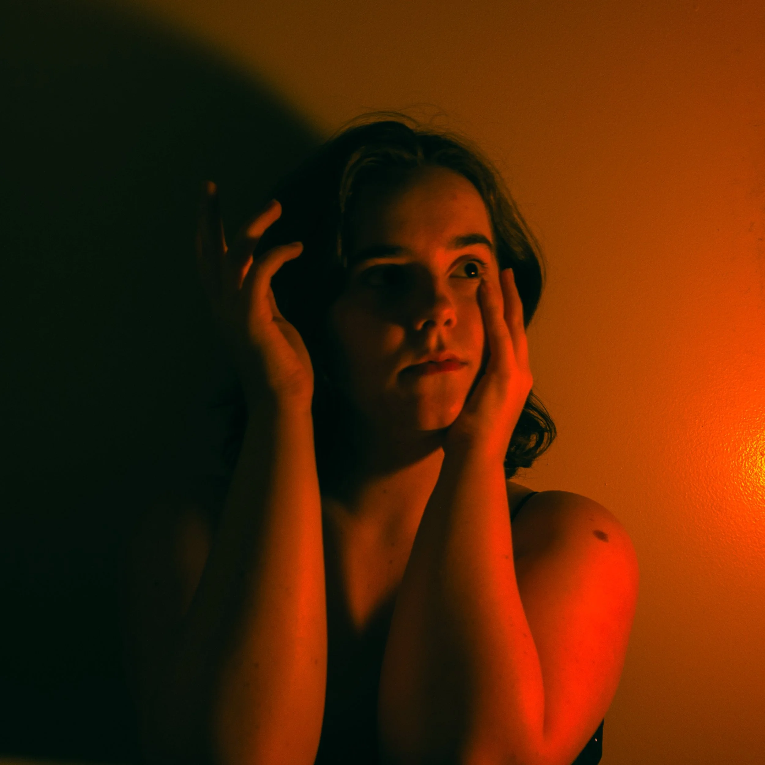 Close-up of a woman's face illuminated by warm orange lighting, holding her head with both hands, with a surprised or contemplative expression.
