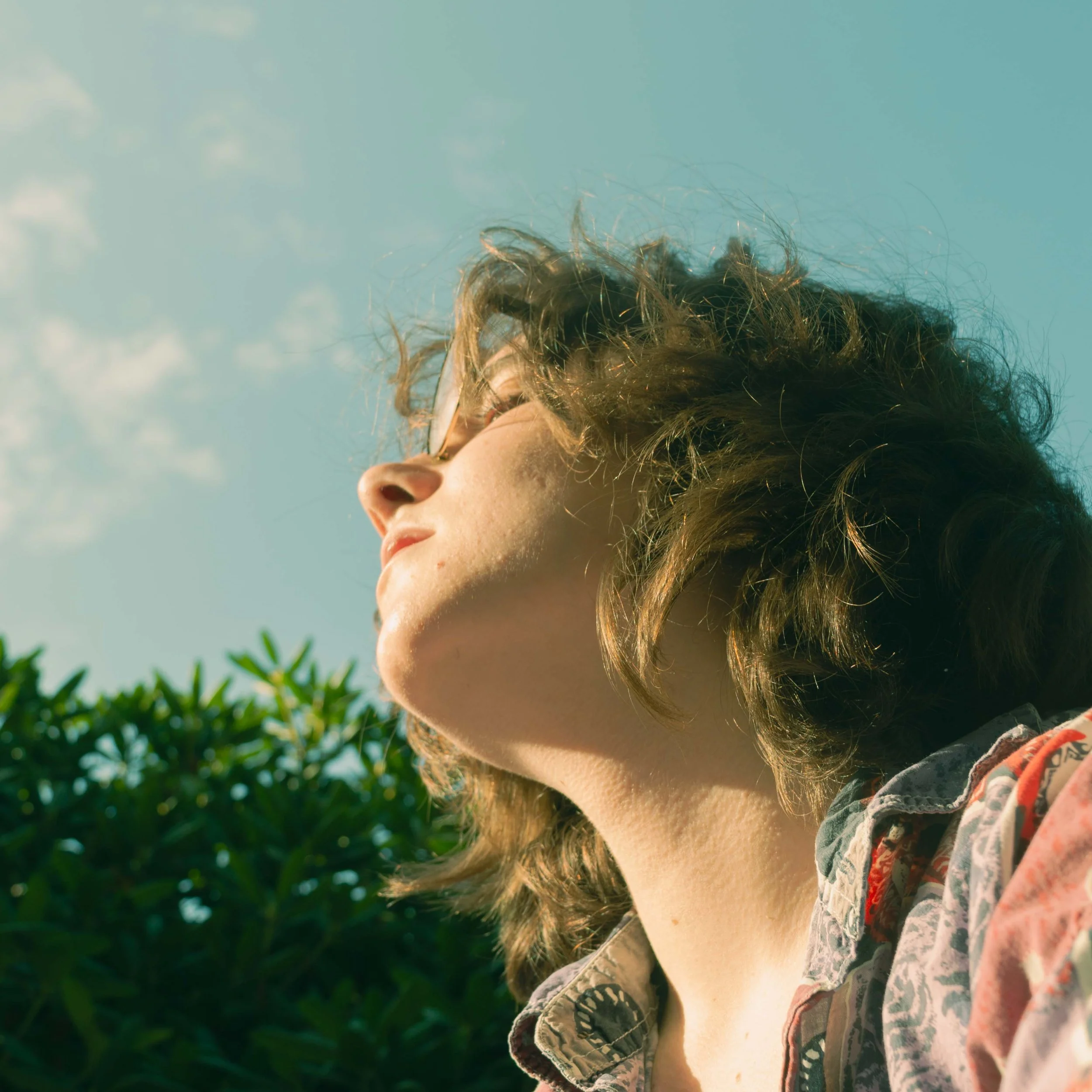 Close-up of a young person with curly hair and glasses, wearing printed clothing, looking towards the sky, with trees and a blue sky in the background.